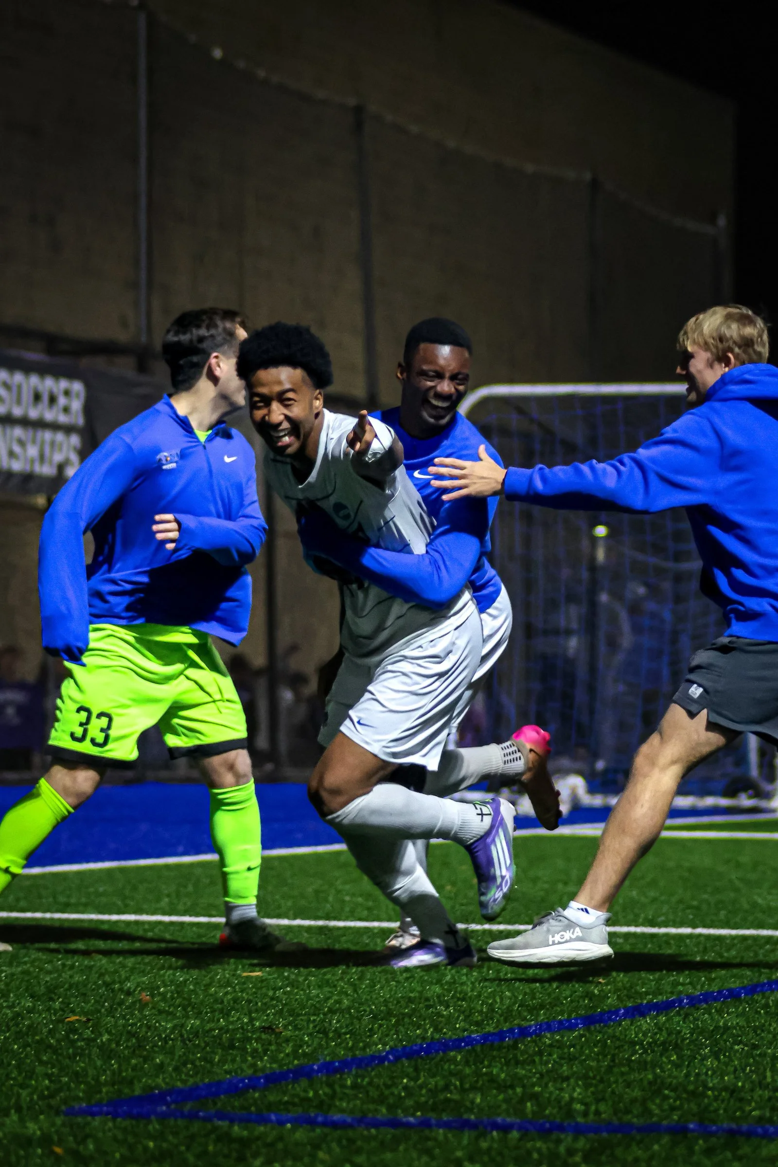 A group of soccer players celebrating on an artificial turf field at night. Four players are visible; one in a white uniform pointing and smiling, others in blue uniforms, one in yellow shorts. They are smiling and laughing together.