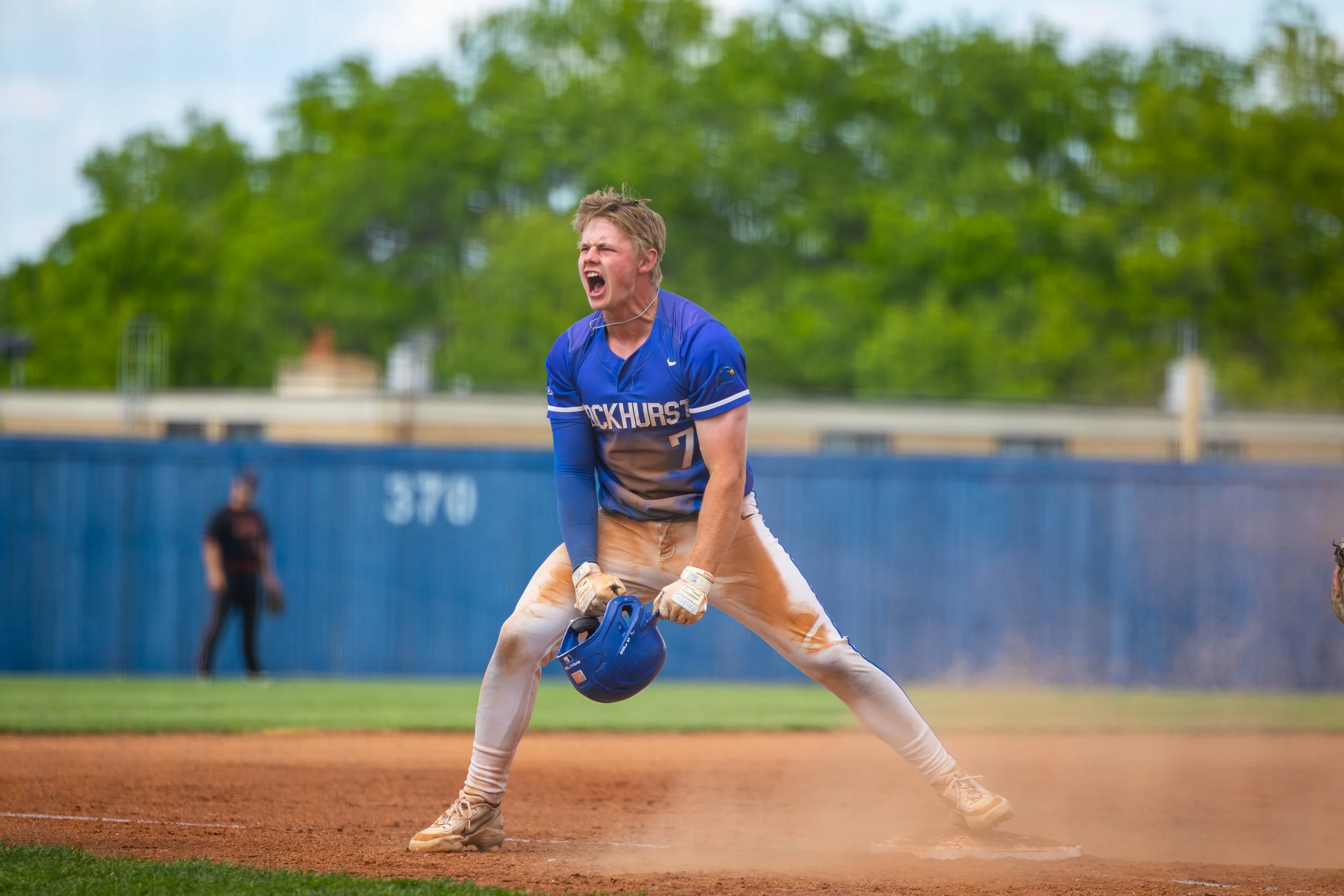 A young male baseball player in a blue and white uniform is at bat, swinging a baseball bat. He is wearing a blue helmet, and has an intense expression with his mouth open. The background shows a baseball field with a blue fence and some trees.