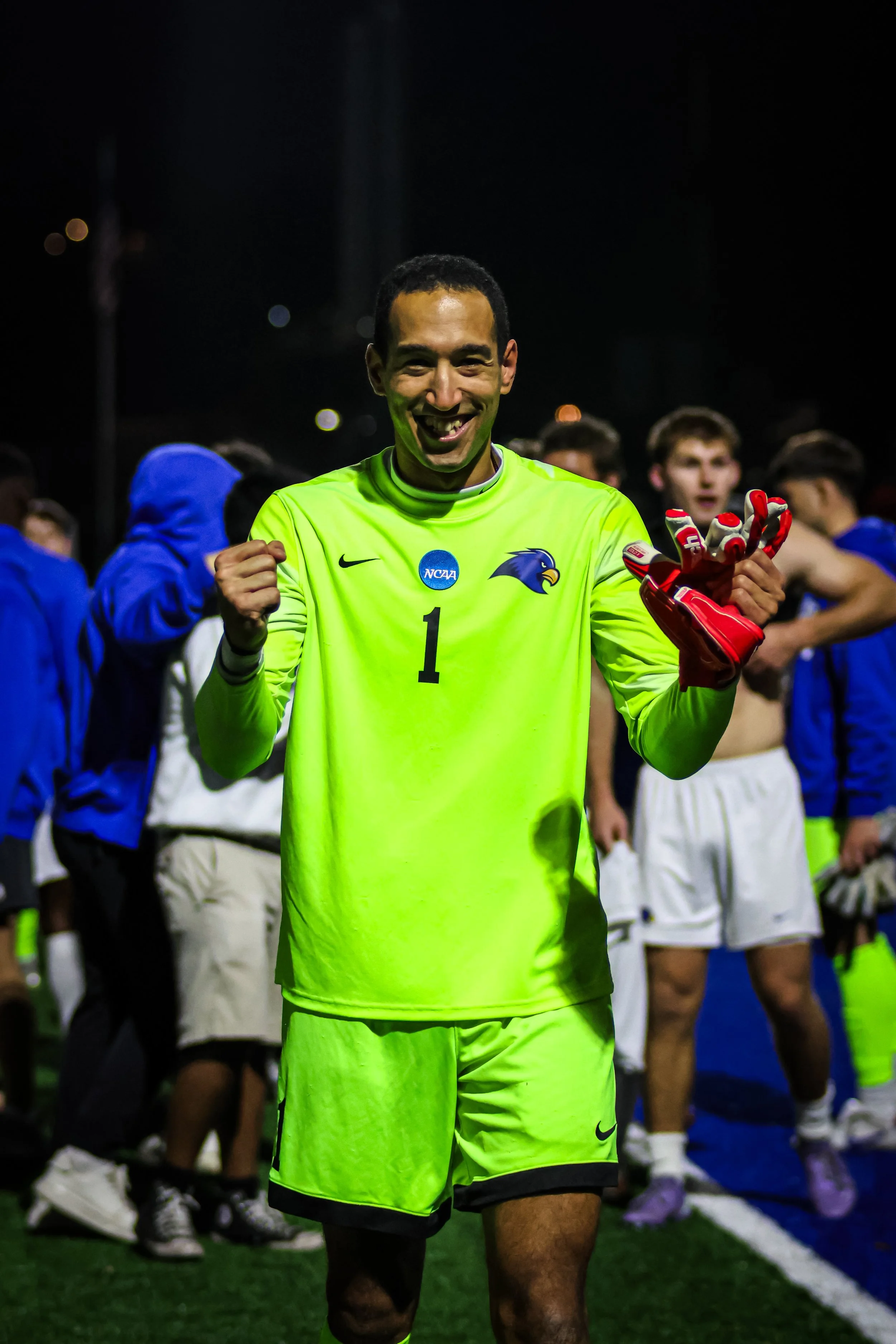 A male soccer goalkeeper celebrating on the field at night, wearing a bright yellow jersey with the number 1, an NC AA patch, and a team logo, holding red goalkeeper gloves with a group of players in the background.