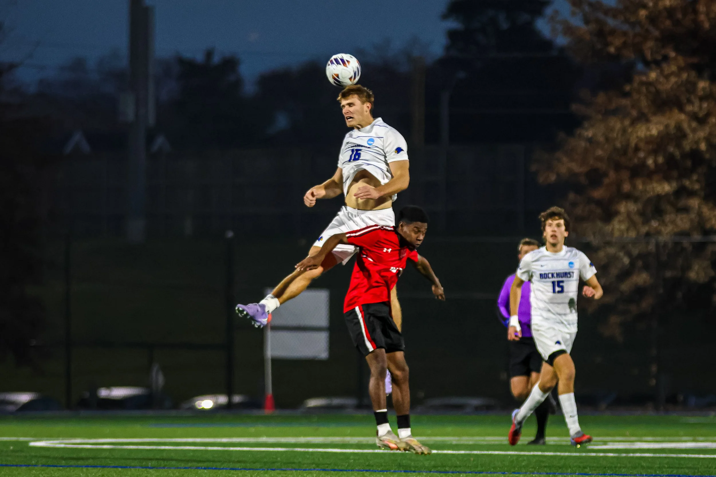 A soccer player wearing a white jersey with the number 16 heads a ball, while another player in red is jumping underneath. Other players and trees are visible in the background.
