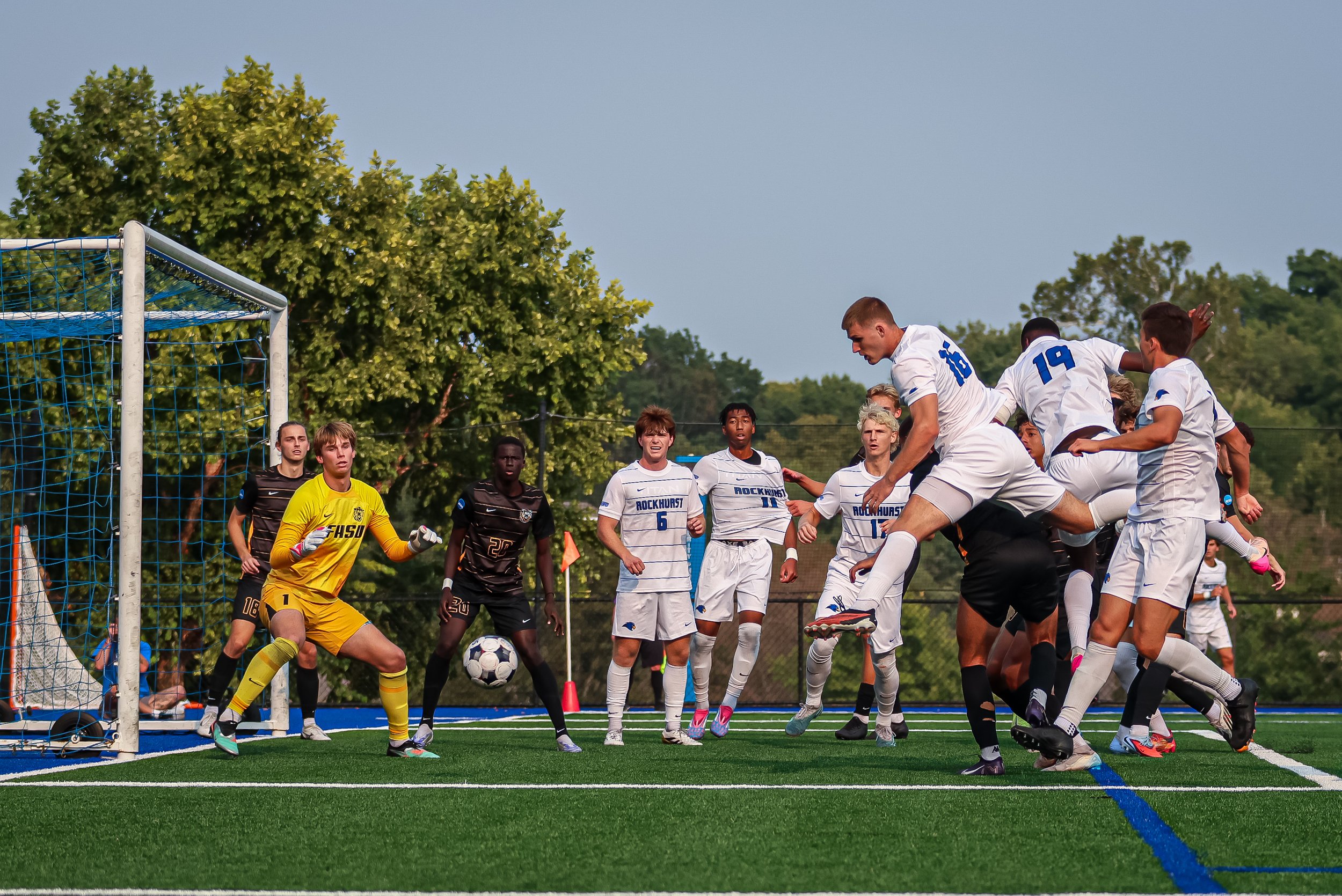A soccer game with players in white and black uniforms, a goalkeeper in yellow, and a ball near the goal, on a field with trees in the background.