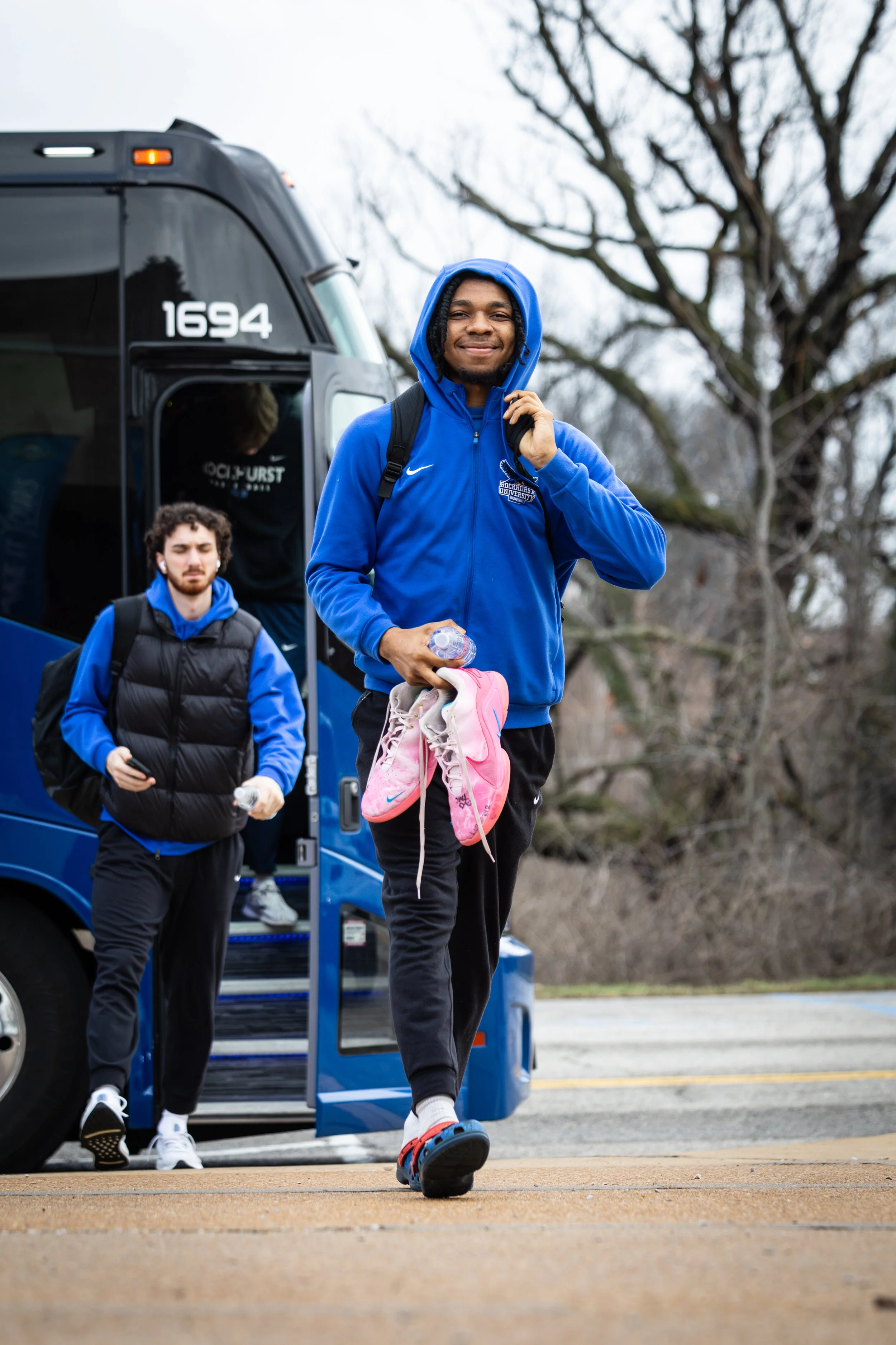 Young man in a blue hoodie and black pants with a pink sneaker in hand, smiling as he steps out of a blue bus. Other individuals also with backpacks exit the bus in a park or outdoor area with leafless trees.