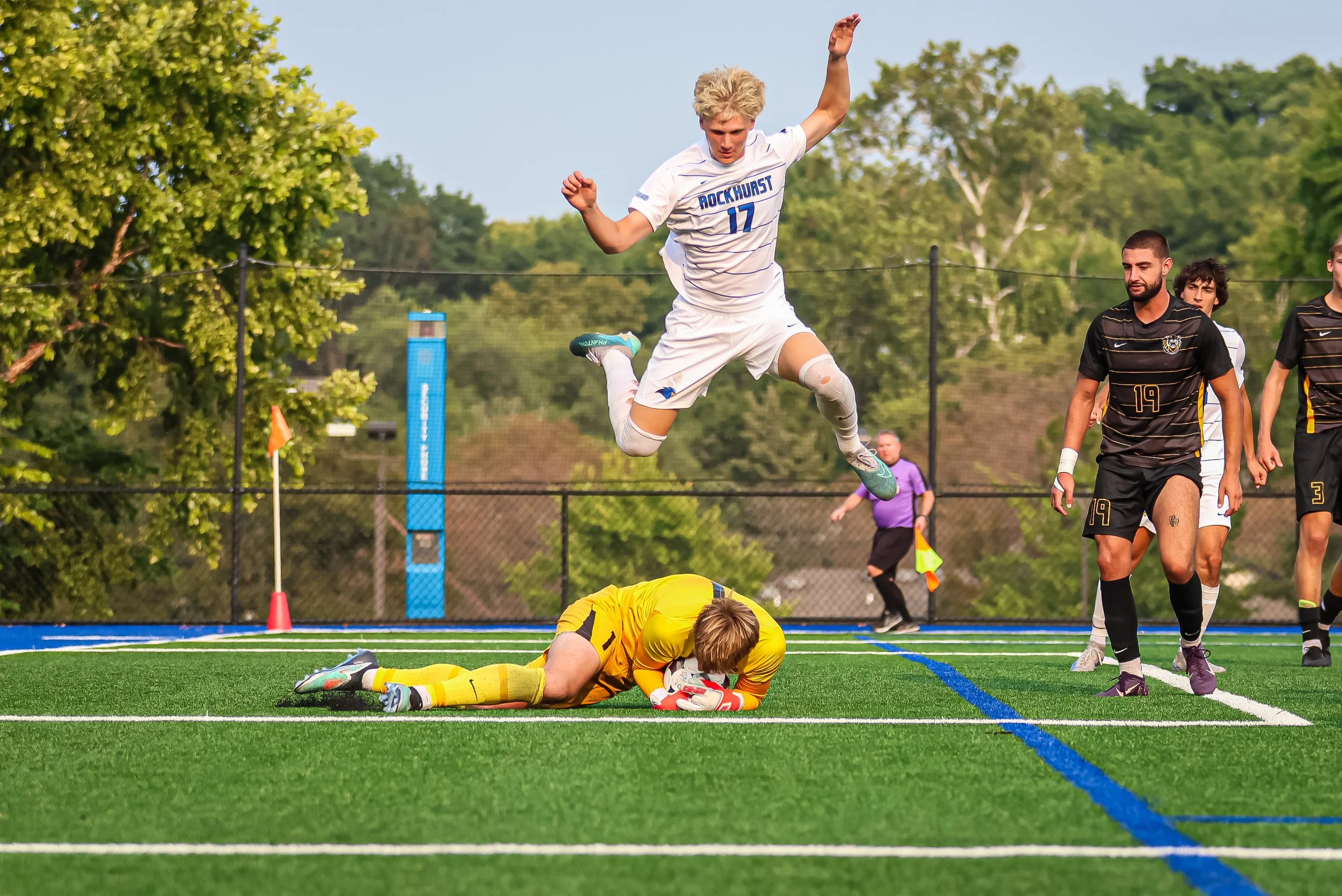 A soccer player in a white uniform jumps to score a goal past a goalkeeper in yellow lying on the ground. Two other players and a referee are observing, with green trees and a blue scoreboard in the background.