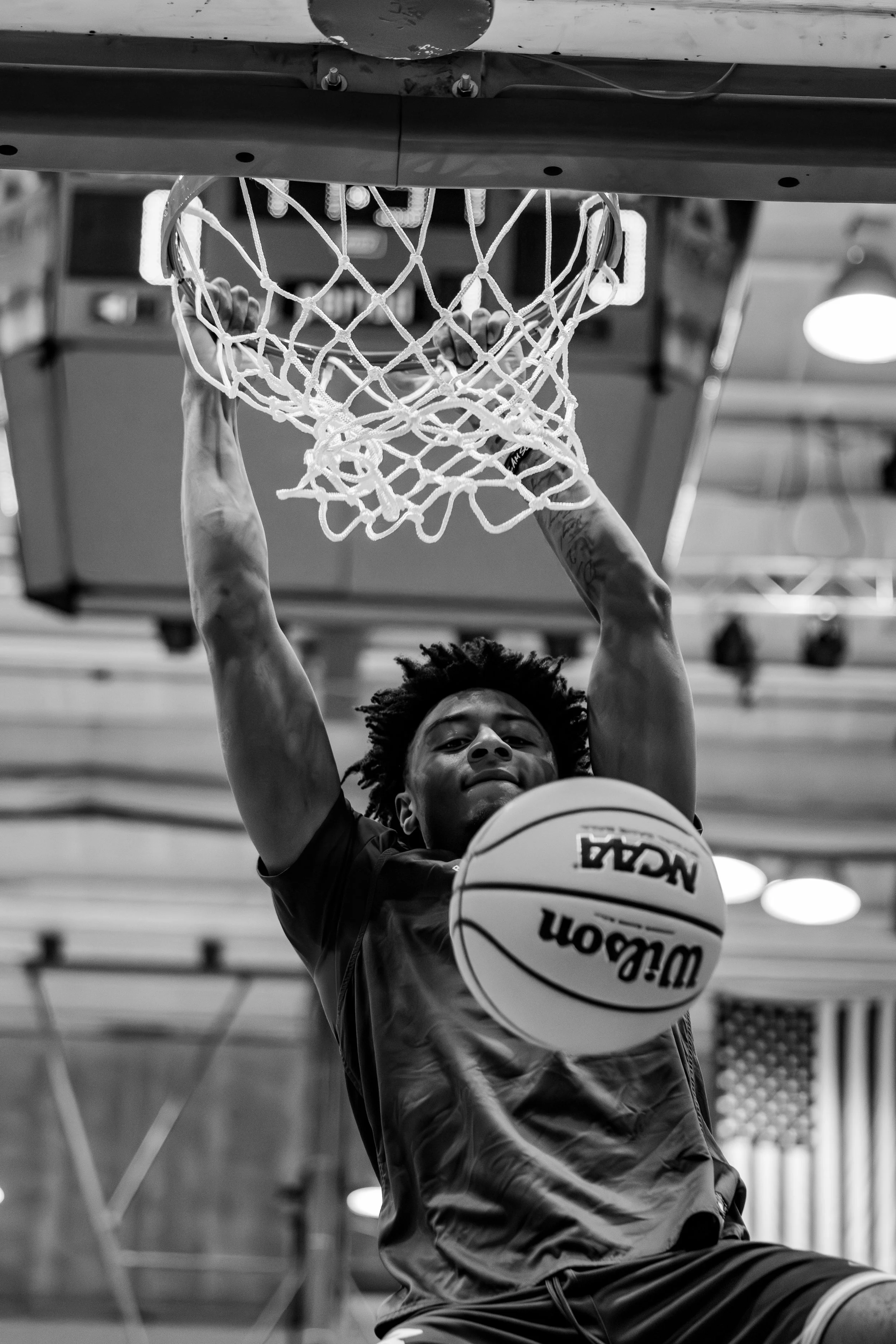 A basketball player is jumping to the basket, holding a basketball in front of him, with both hands, while hanging from the rim with his other hands, in an indoor gym.