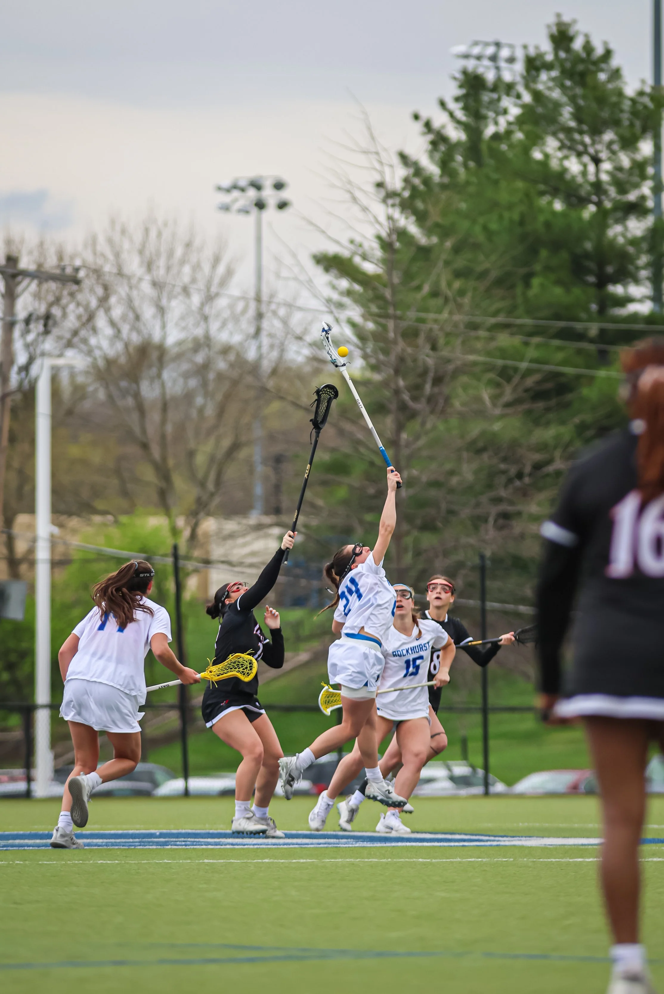 Girls playing lacrosse on a field with trees and light poles in the background, reaching up to catch or block a yellow ball.