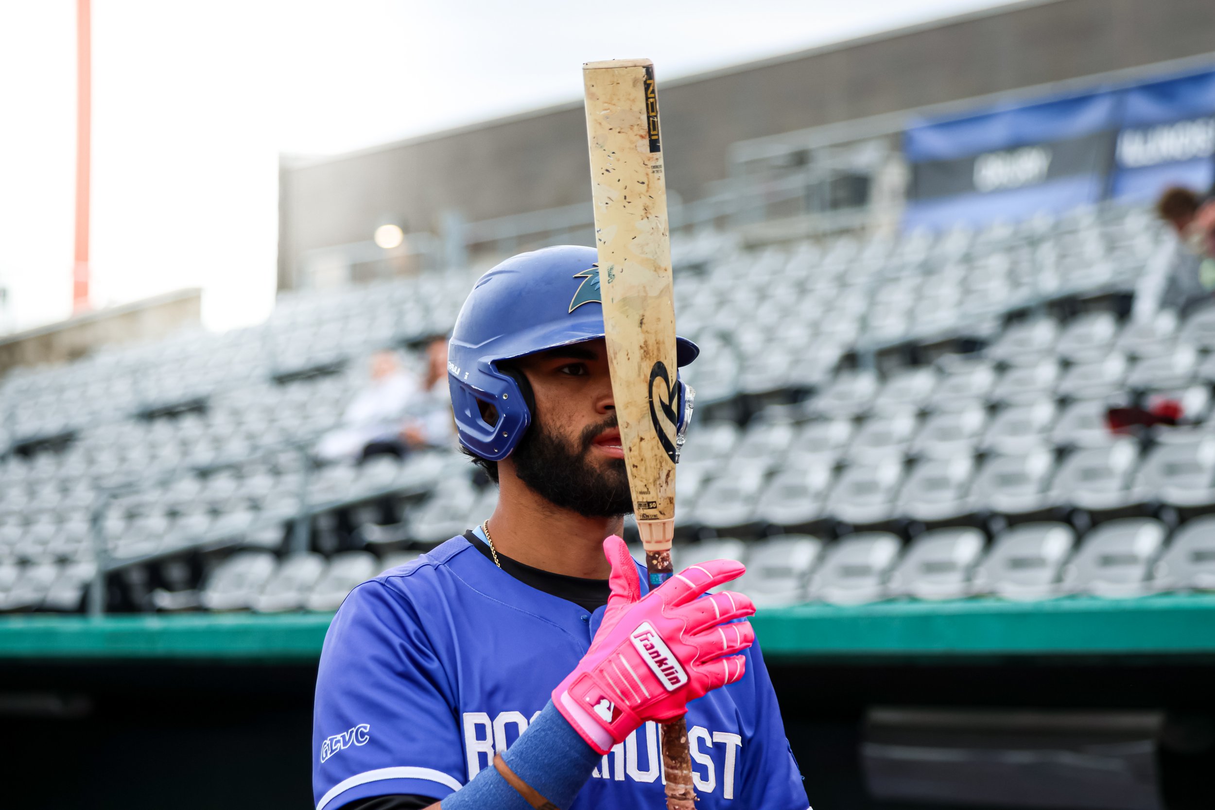 A man wearing a blue baseball helmet and matching shirt, holding a baseball bat vertically in front of his face, with a bright pink glove visible on his hand at a sports stadium.