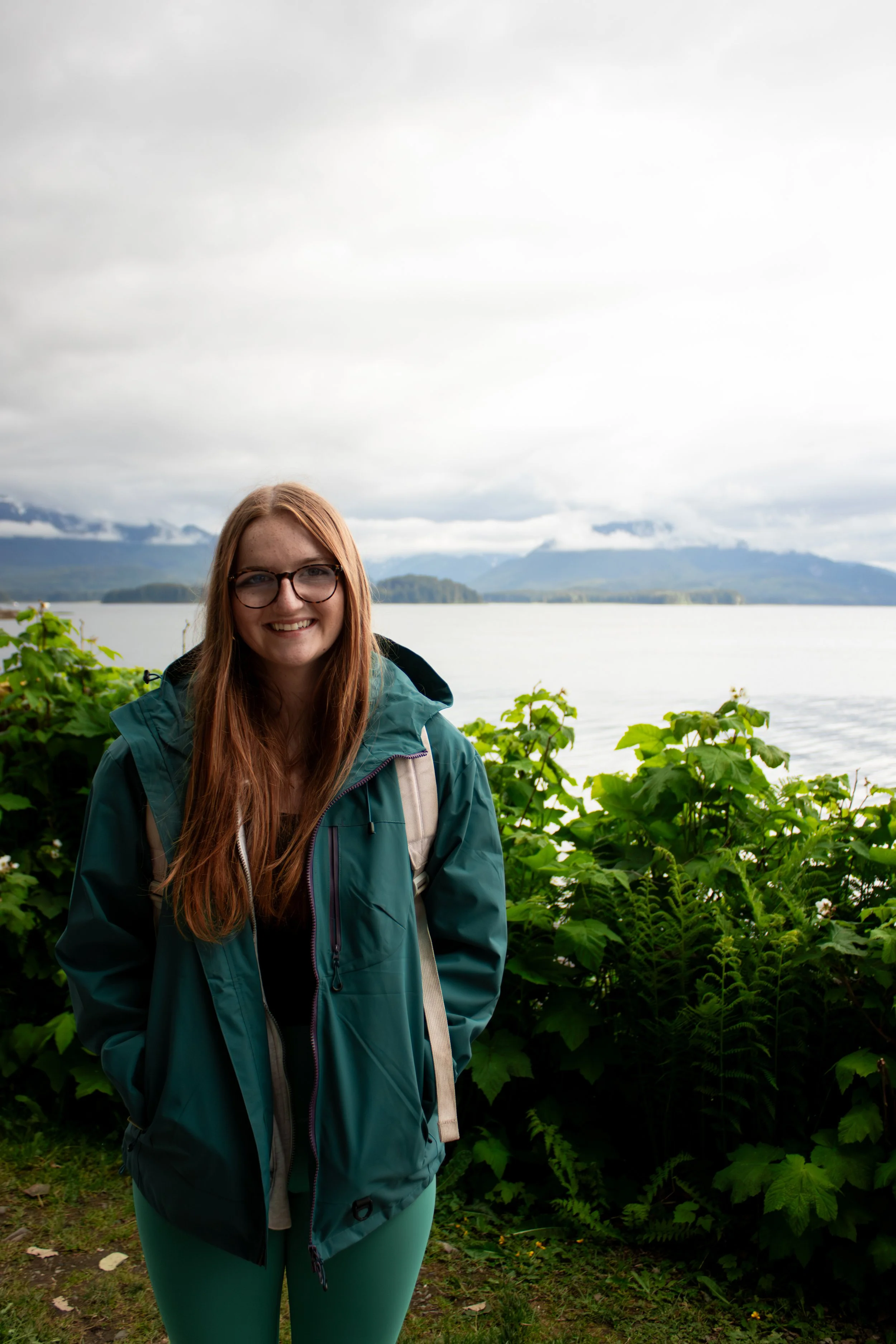A smiling woman with long red hair and glasses standing outdoors near a lake or river with green plants in the foreground and mountains in the background, overcast sky.