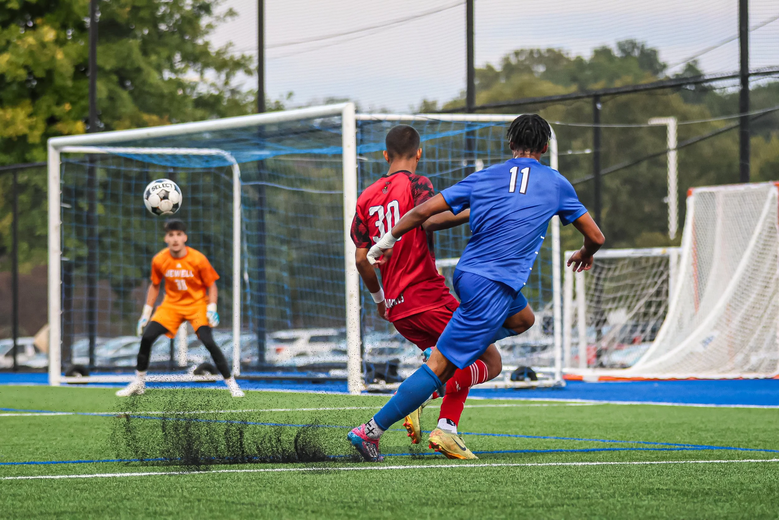 Two soccer players, one in a red uniform and the other in a blue uniform, are competing for the ball near the goal. The goalkeeper in an orange uniform is watching the play unfold behind them, with a soccer net and trees in the background.