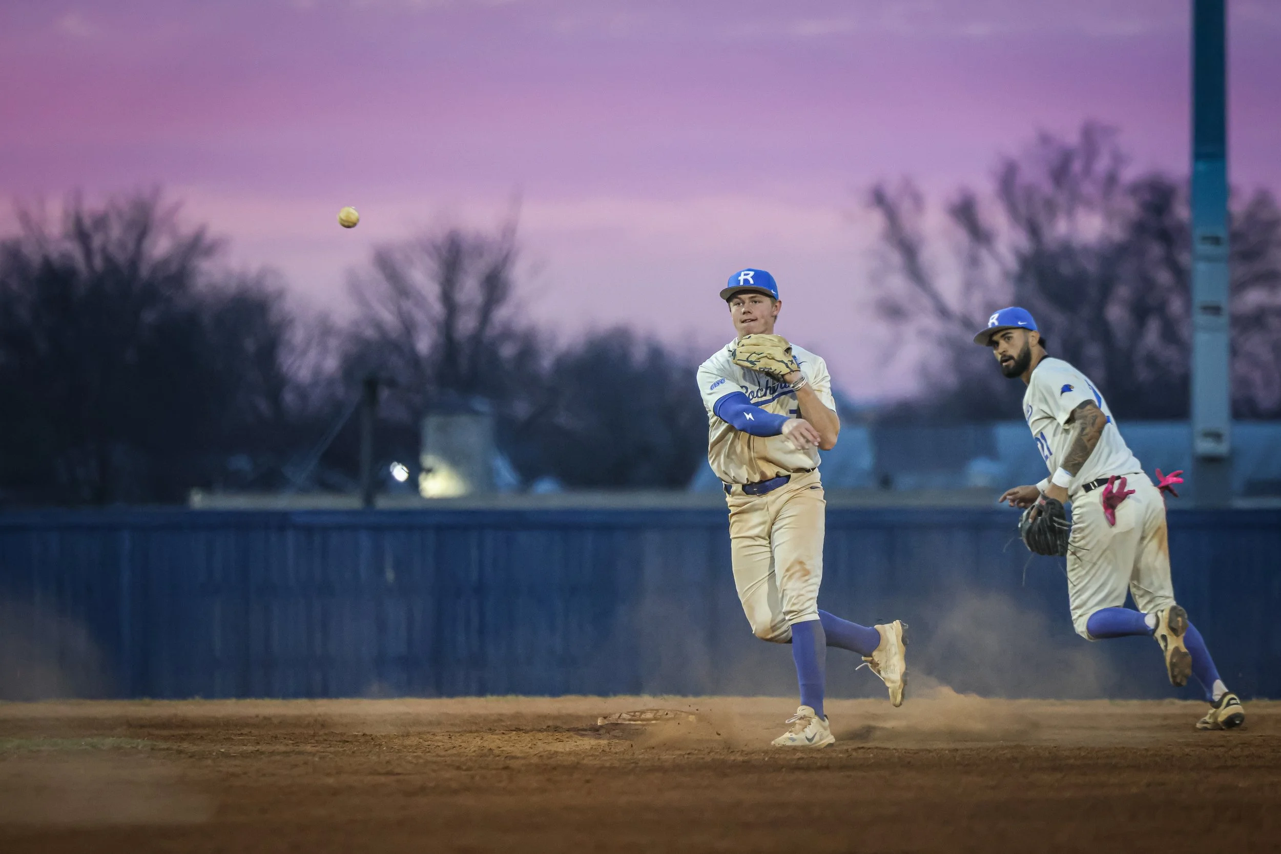 Two baseball players, one in the middle and one on the right, are on a baseball field during dusk. The player in the middle is preparing to catch a ball that is in the air, while the player on the right is looking towards the ball. Both are wearing b