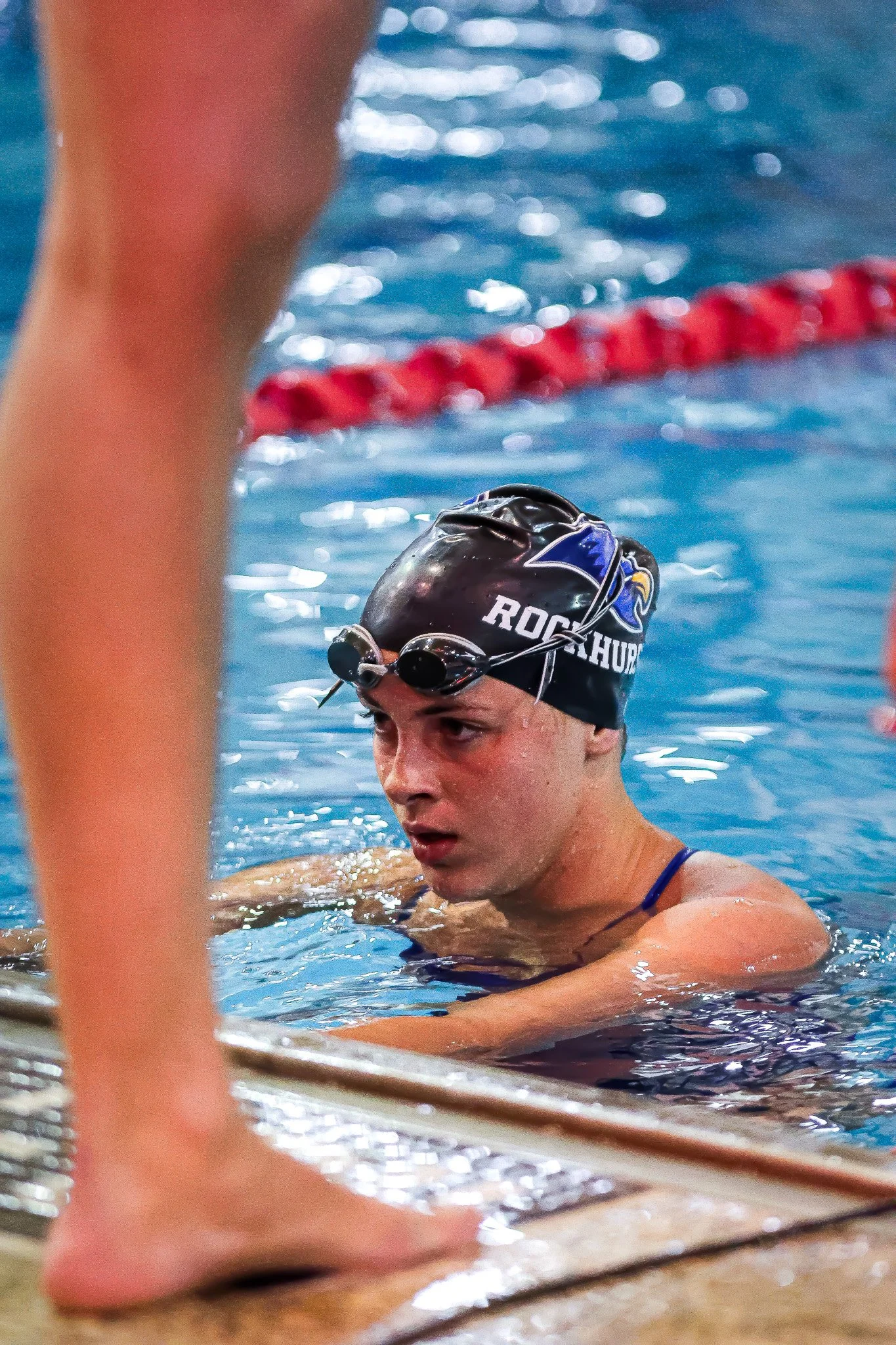 A male swimmer wearing a black swim cap with the words 'Rockin' and a logo, and goggles, resting at the edge of a swimming pool during a competition or practice.