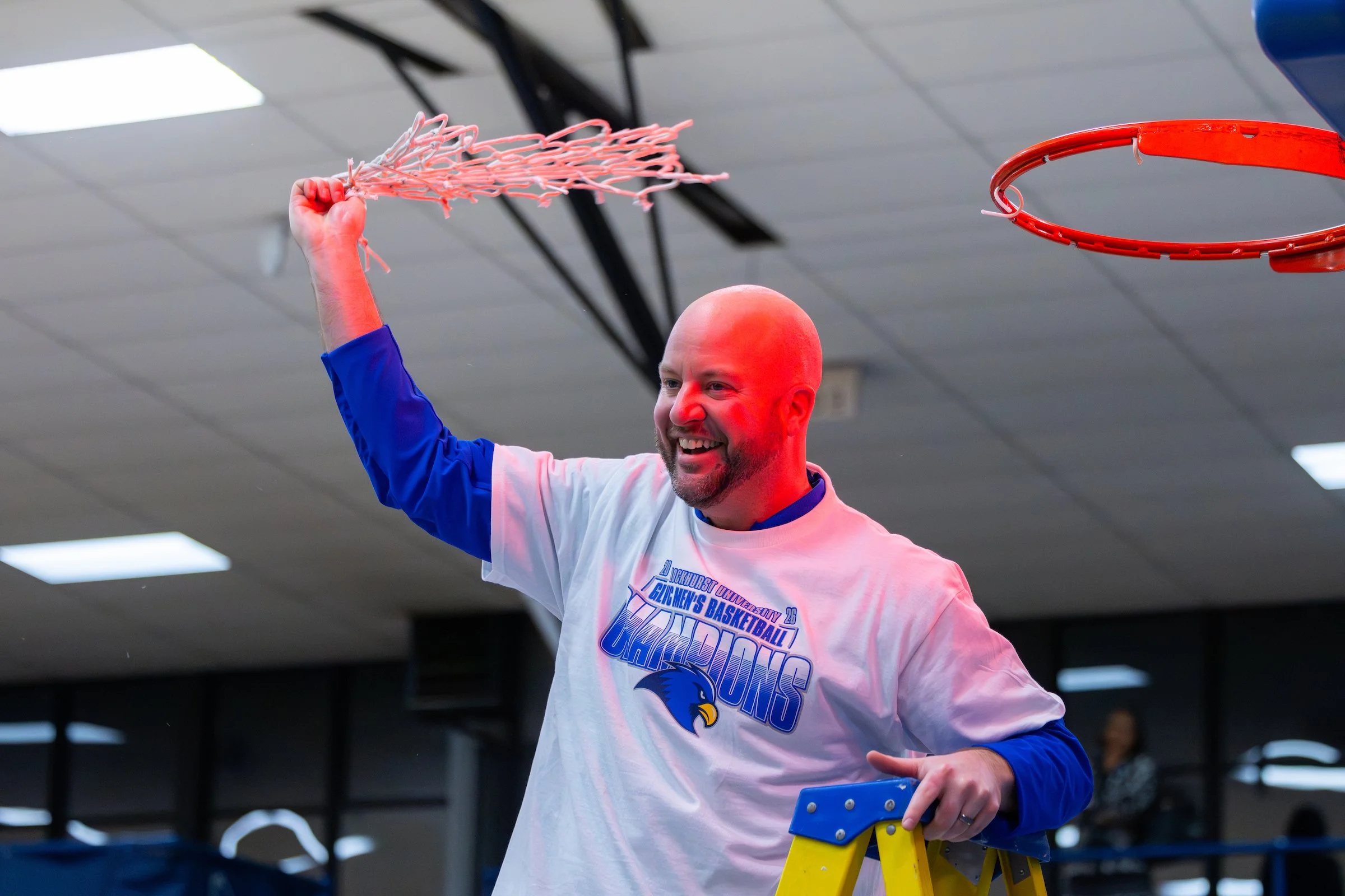 A man with a bald head and a beard smiles as he raises a basketball hoop net with his right hand. He is wearing a white and blue basketball jersey with the logo of a university team. The background shows a ceiling with fluorescent lights and black gy