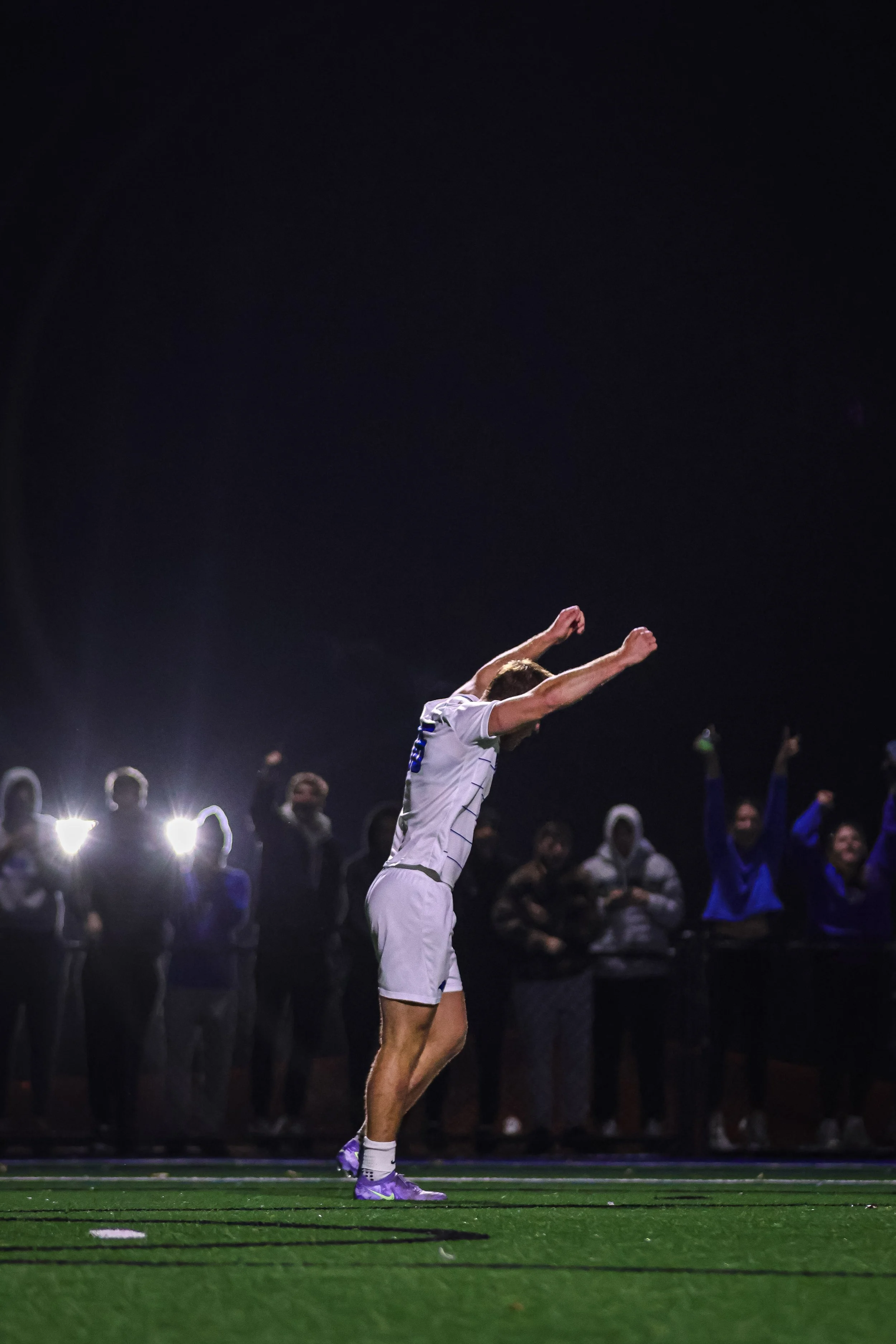 A soccer player wearing a white uniform is celebrating with arms raised on a green turf field at night, with spectators in the background.