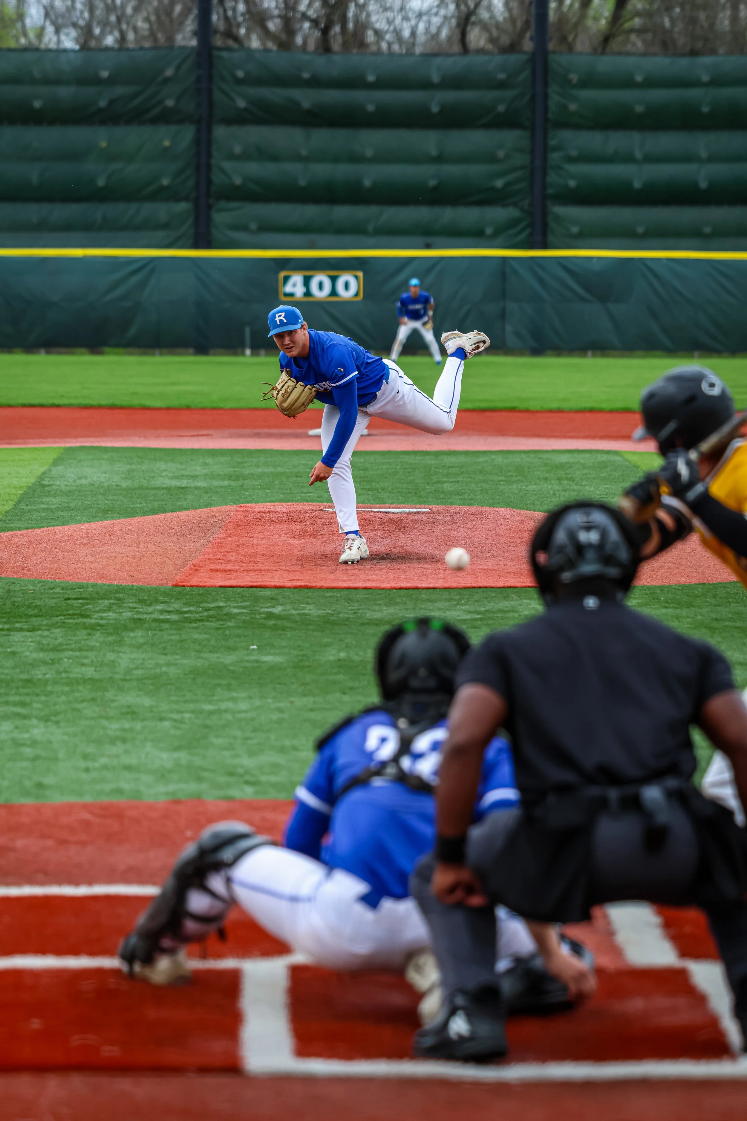 A baseball pitcher in a blue uniform throws a pitch during a game on a field with green grass and a scoreboard in the background. The catcher is positioned behind home plate wearing a black helmet and gear, and two players are seated behind the catch