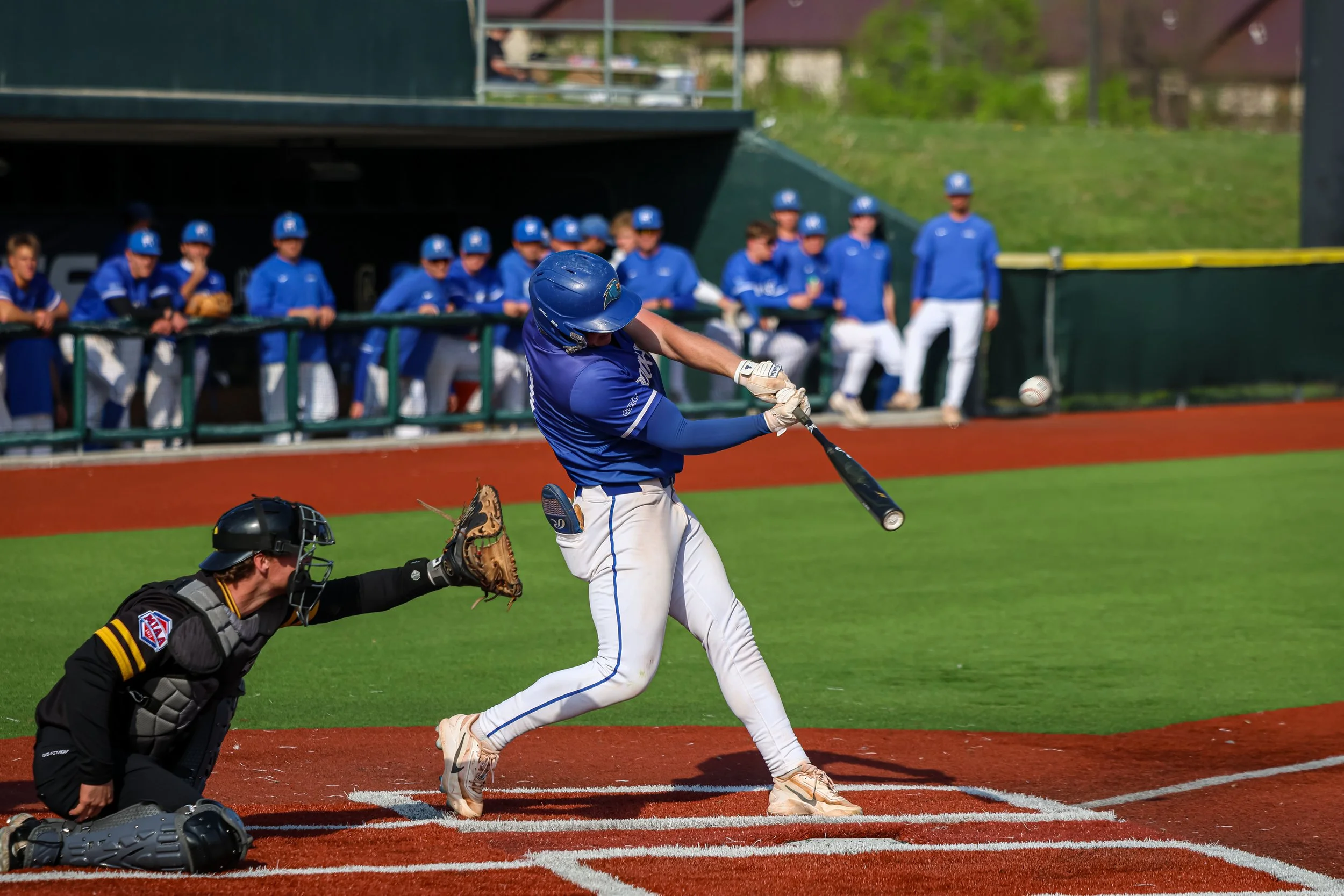 A baseball player in a blue uniform is swinging at a pitch at home plate during a game. The catcher is crouched behind him and an umpire is observing. Several players in blue uniforms are in the dugout watching the game.