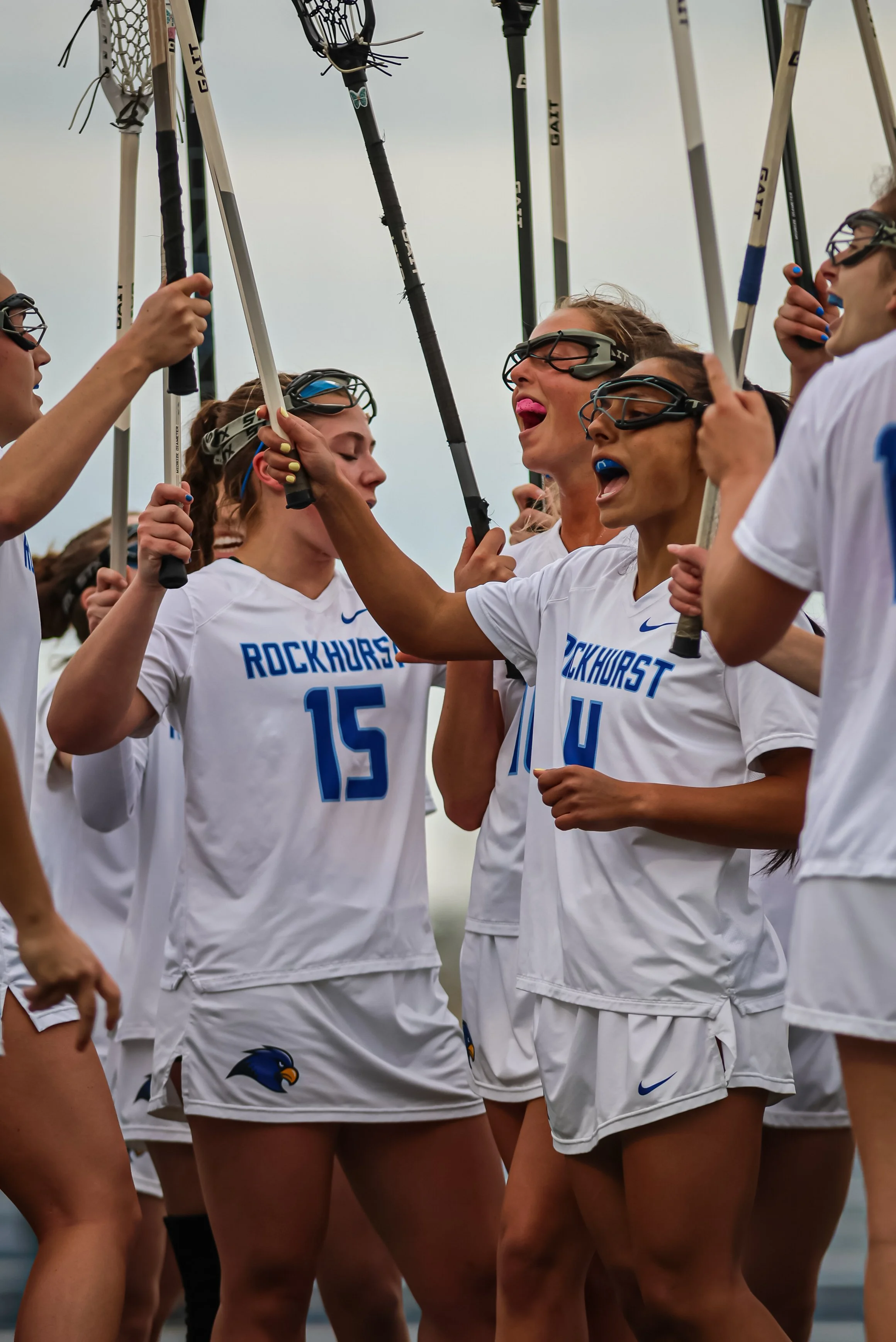 Women wearing white sports uniforms with 'Rockhurst' written on the front, holding lacrosse sticks, cheer and celebrate during a game or practice on an overcast day.