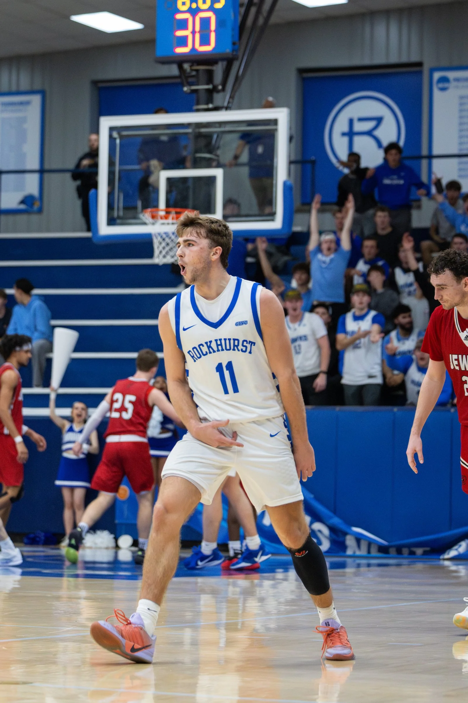A basketball player in a white jersey with blue accents, number 11, from Rockhurst, is on the court with a focused expression. Other players, spectators, and a digital scoreboard are visible in the background of a gymnasium.