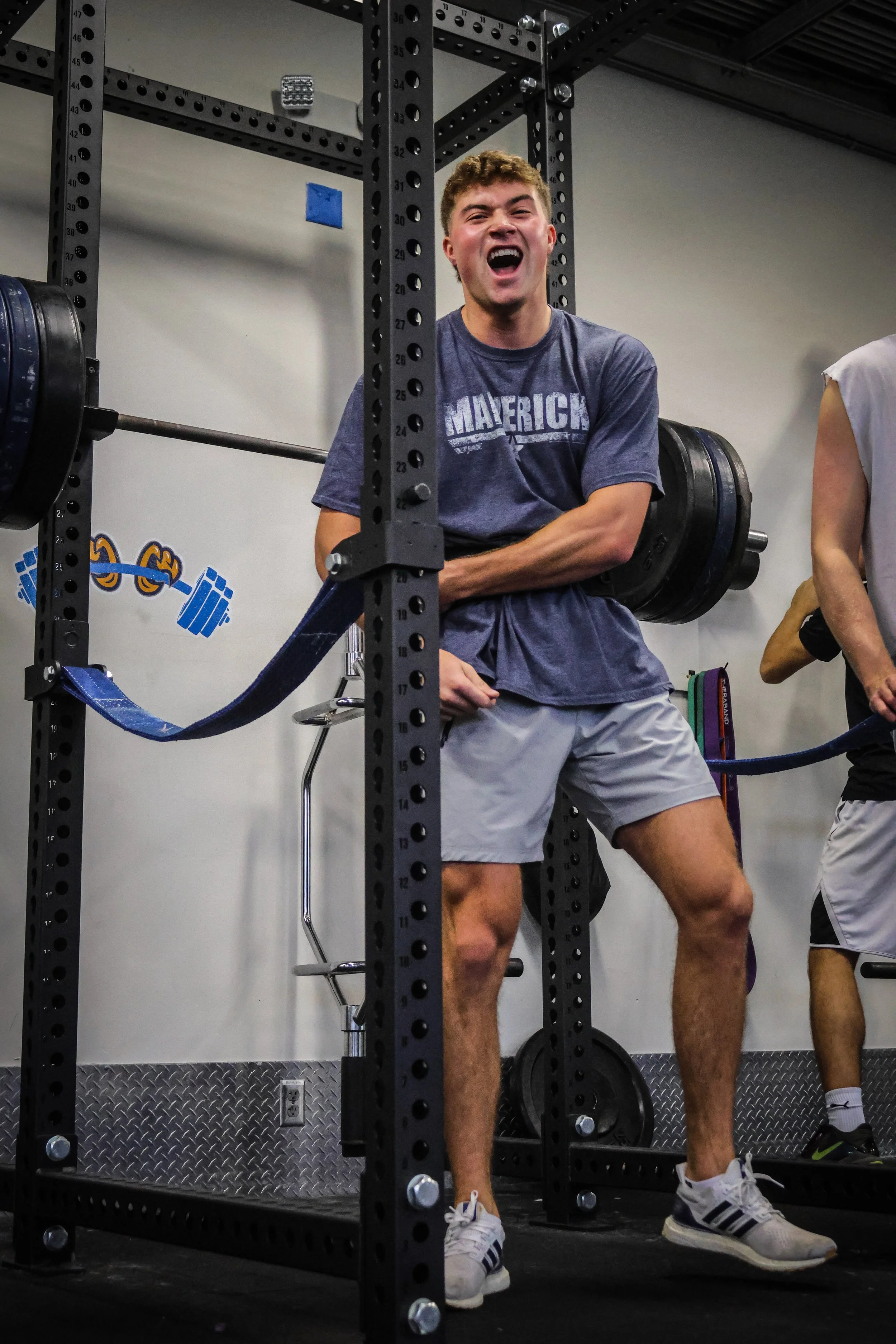 A young man with curly hair wearing a navy blue T-shirt and gray shorts, sitting on a weightlifting bench with a barbell loaded with weight plates behind him, mid-laugh in a gym setting with black metal equipment and a blue wall decal.