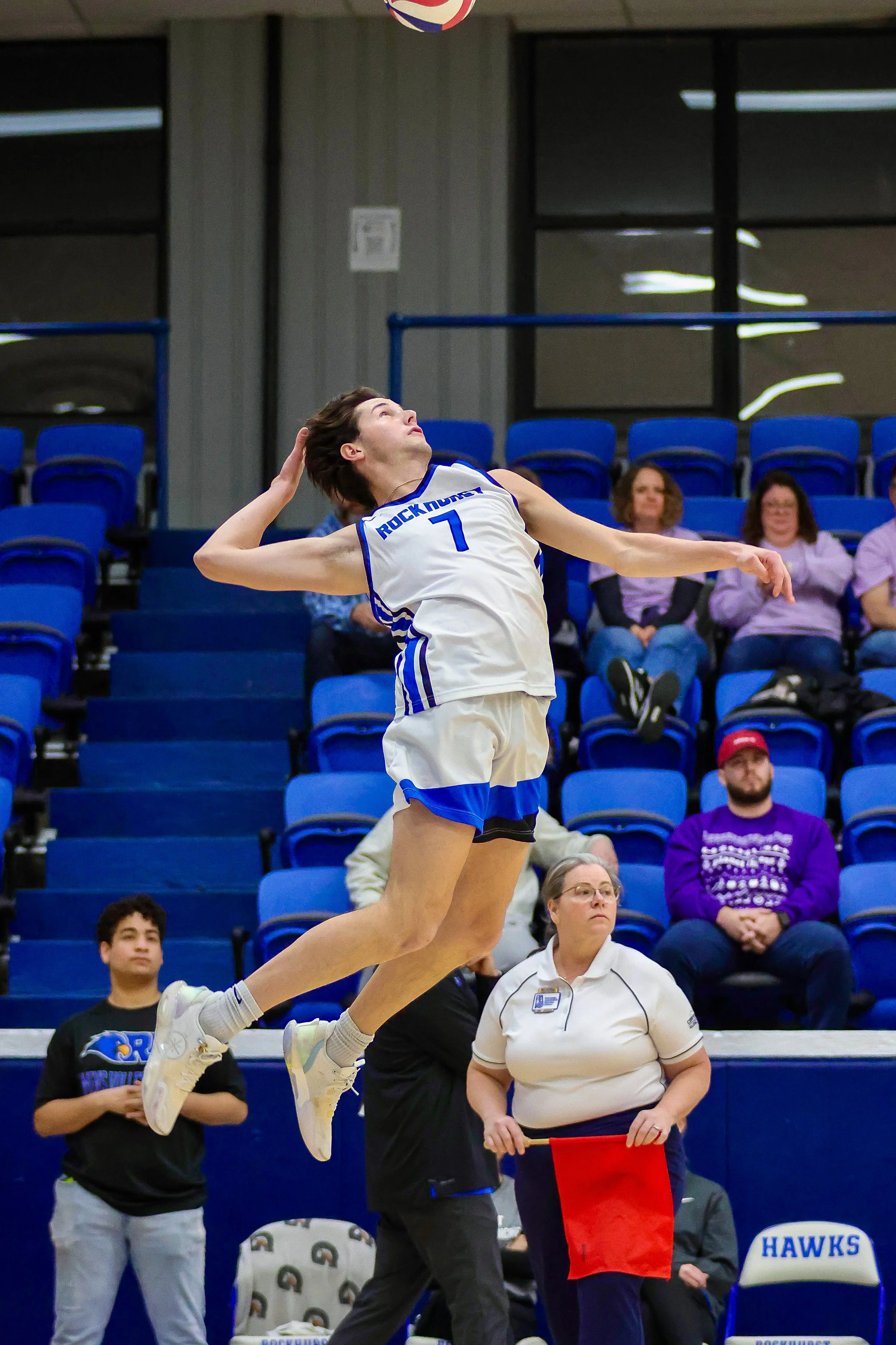 A female volleyball player wearing a white and blue uniform with the number 7 jumps in the air to hit the volleyball. She is on an indoor court with spectators sitting in blue seats and officials present.