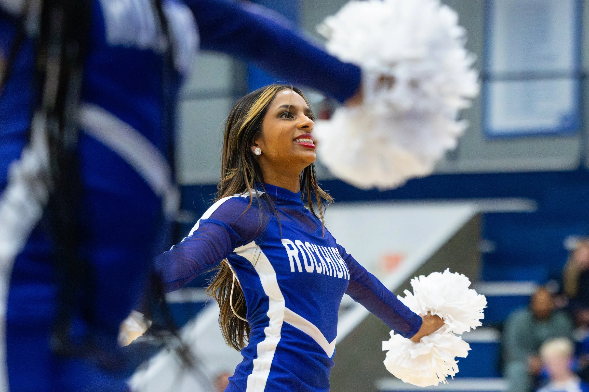 A cheerleader in a blue and white uniform with the word 'Rockies' on it, holding white pom-poms, smiling, during a game or event inside a sports arena.