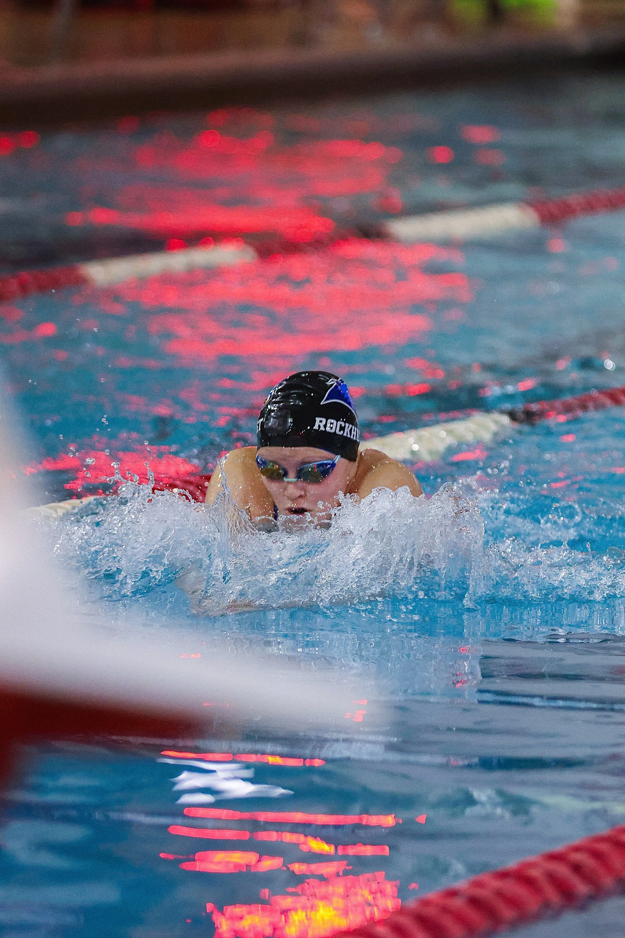 Swimmer in a pool during a competition wearing a black swim cap and goggles, with red lane markers and reflections of red and yellow lights on the water.