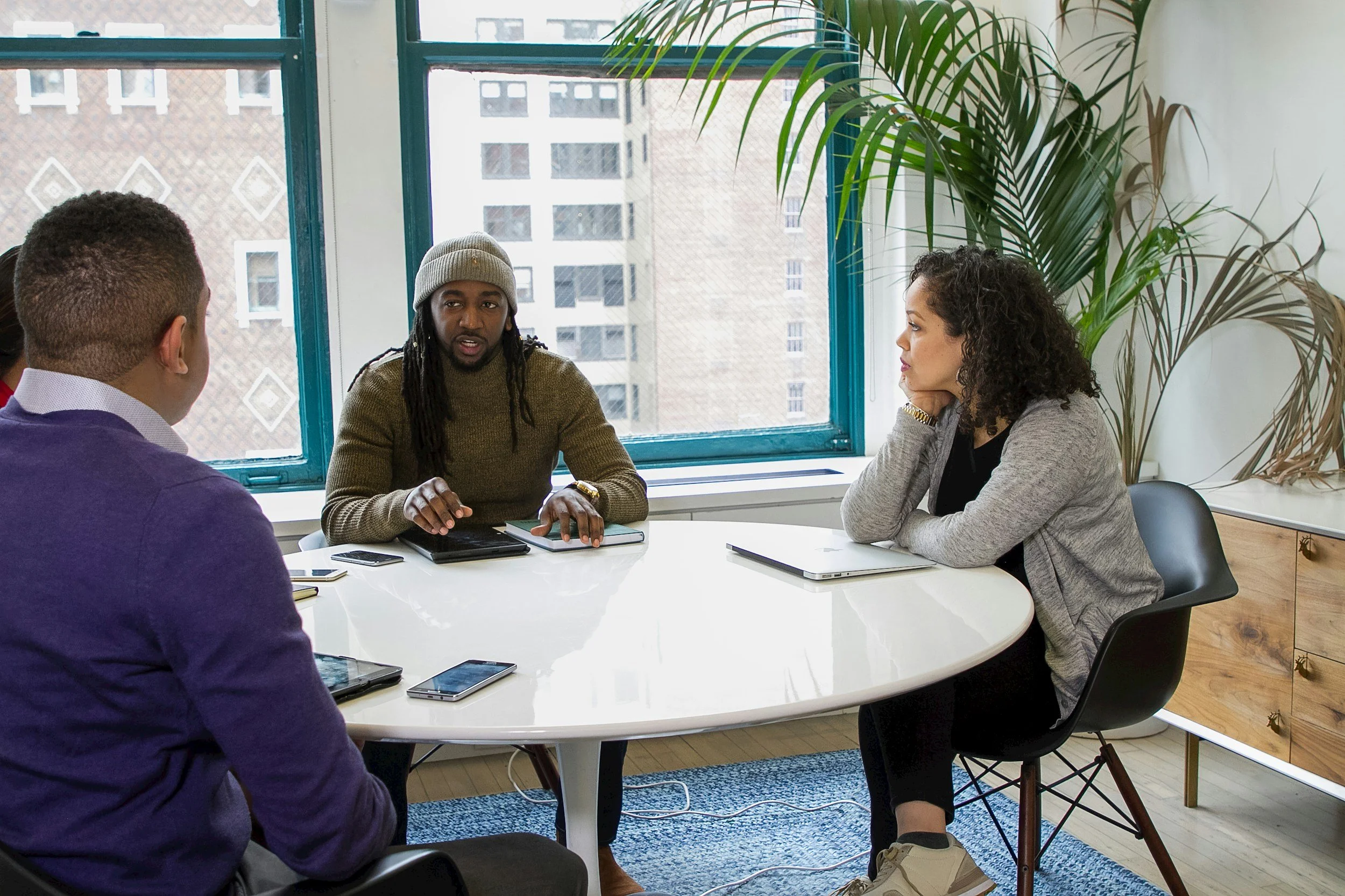 Four people sitting around a white table in a bright room with large windows, engaged in a discussion. One man with dreadlocks and a gray beanie speaking, a woman with curly hair listening attentively, and two men with short hair, partially visible.