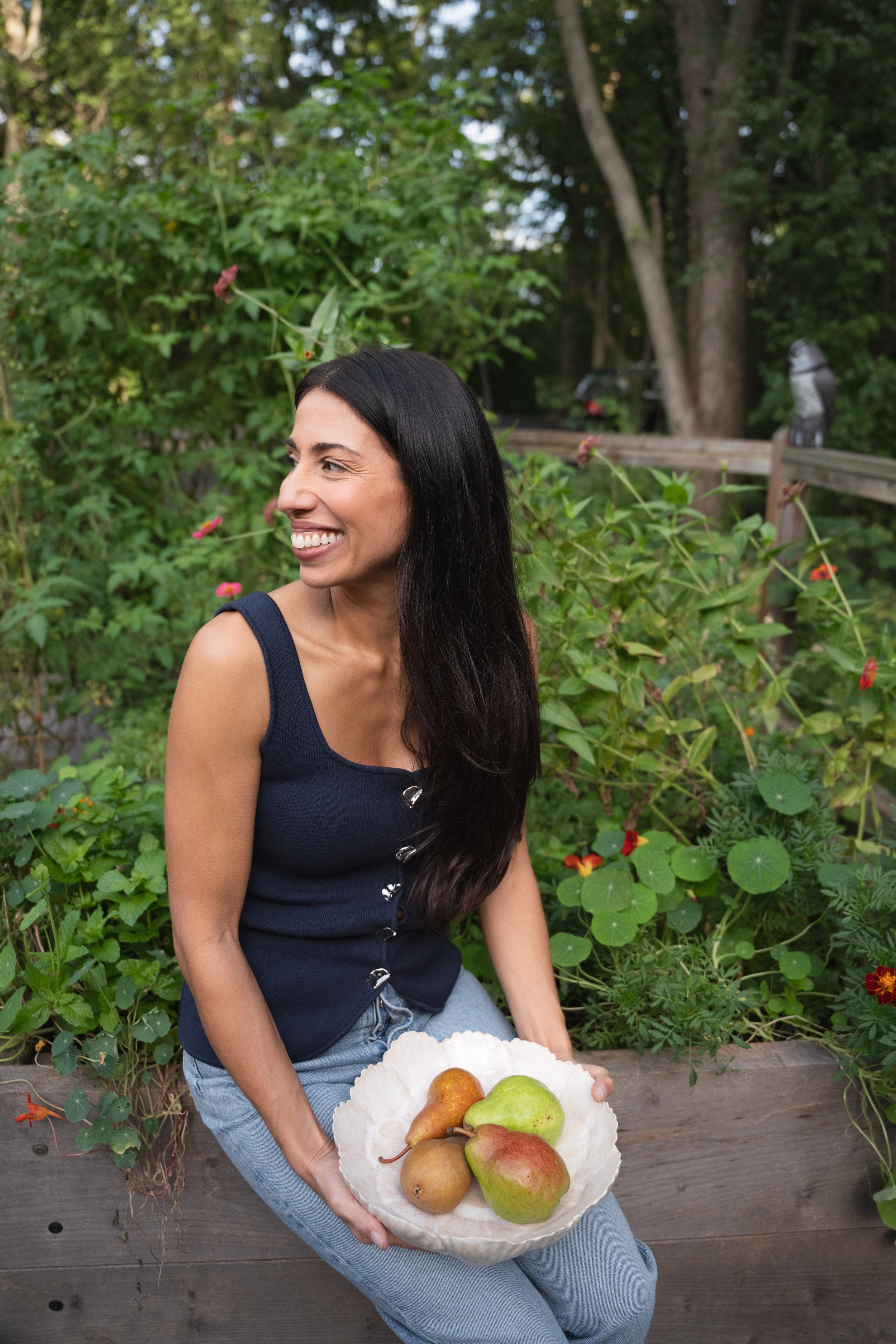Woman sitting outdoors on a wooden bench, holding a white bowl filled with apples, pears, and a pear at a garden with green leafy plants and trees.
