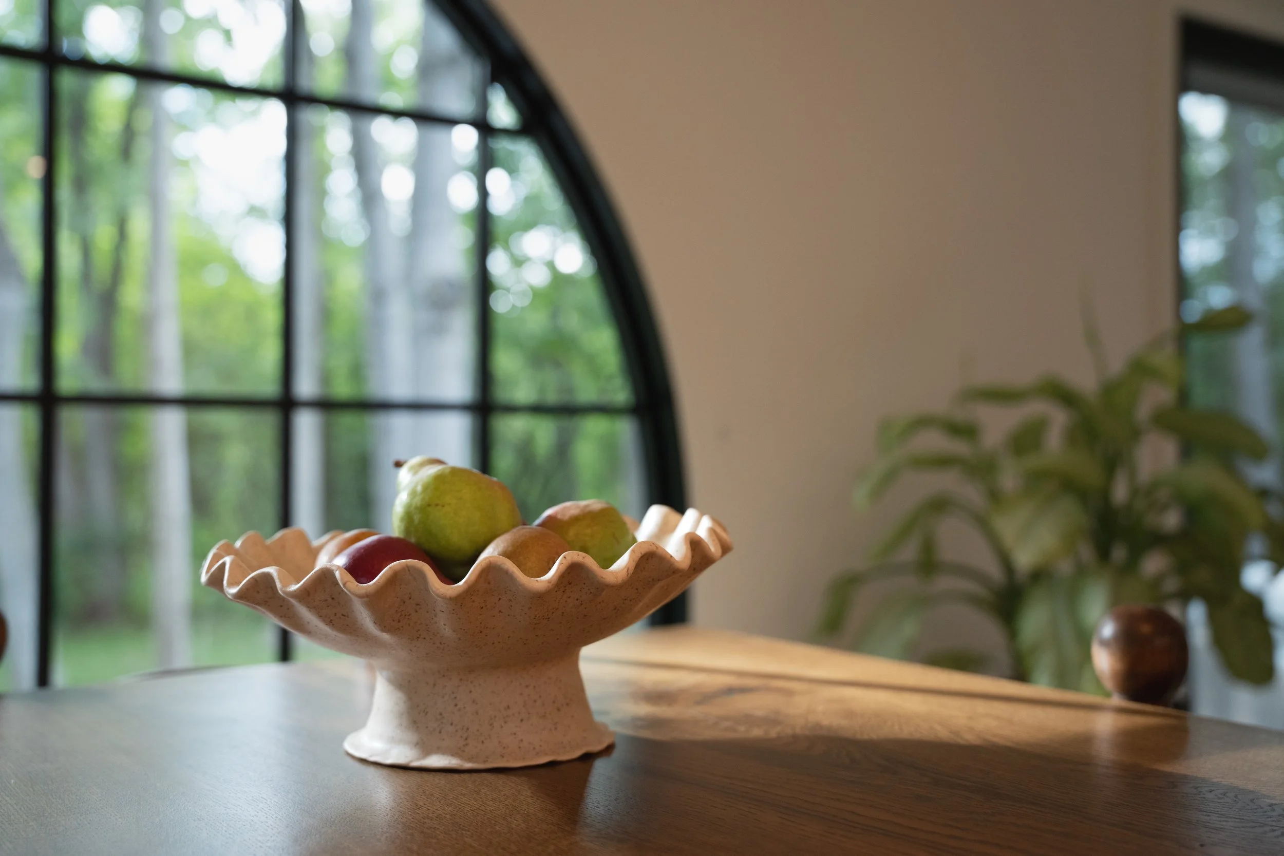 A handmade ceramic pedestal bowl with ruffled edge filled with assorted fresh pears, placed on a wooden table near a window with lush greenery outside.