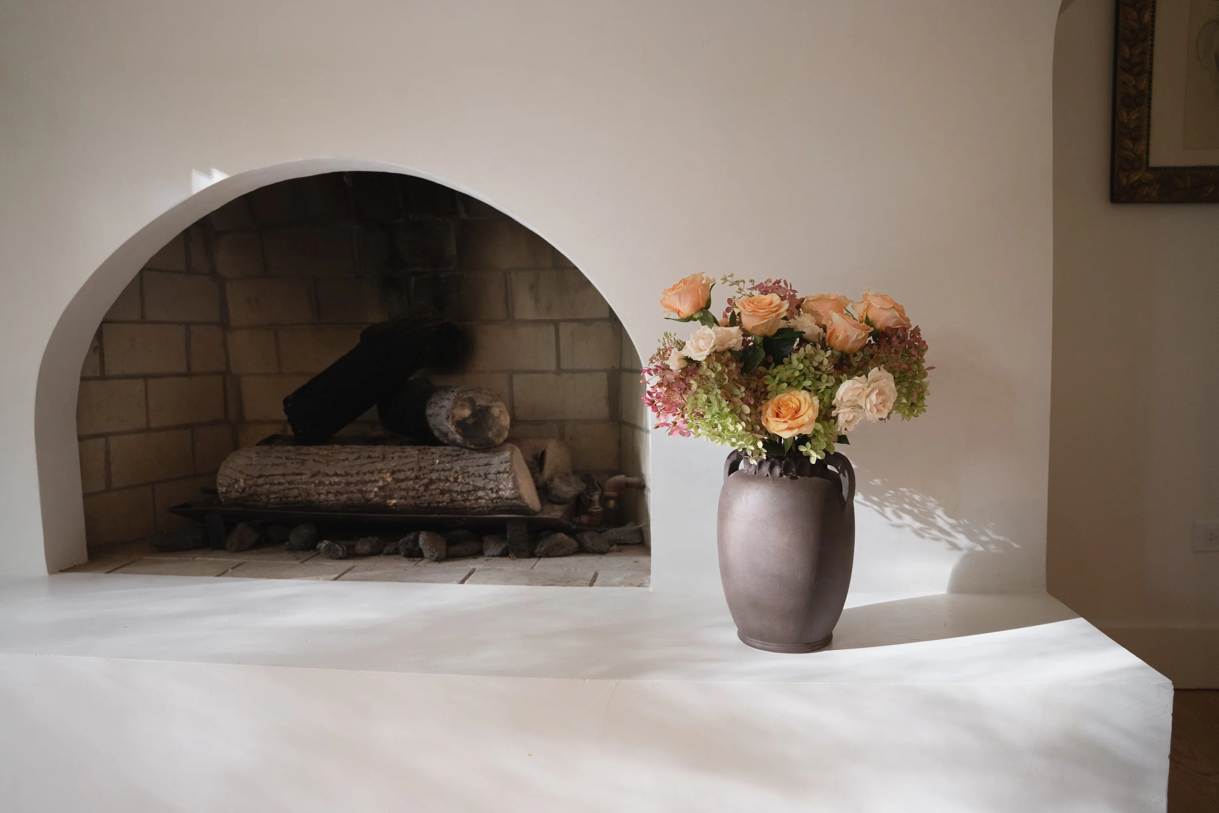 A large handmade chocolate brown vase holding a bouquet of peach and white roses with green and pink foliage, placed on a white surface in front of a white plaster fireplace with logs and brick interior, against a creamy wall.