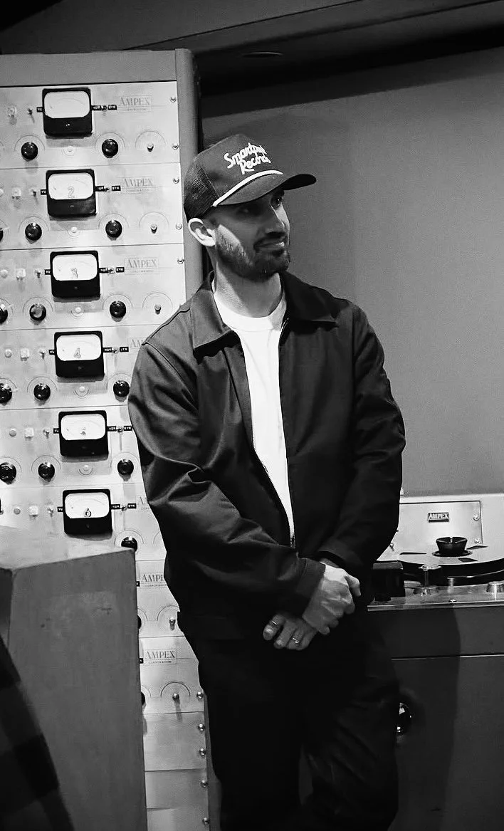 A man standing in a room with audio equipment, wearing a cap with the text 'Sneaky Pete,' a white shirt, and a jacket.