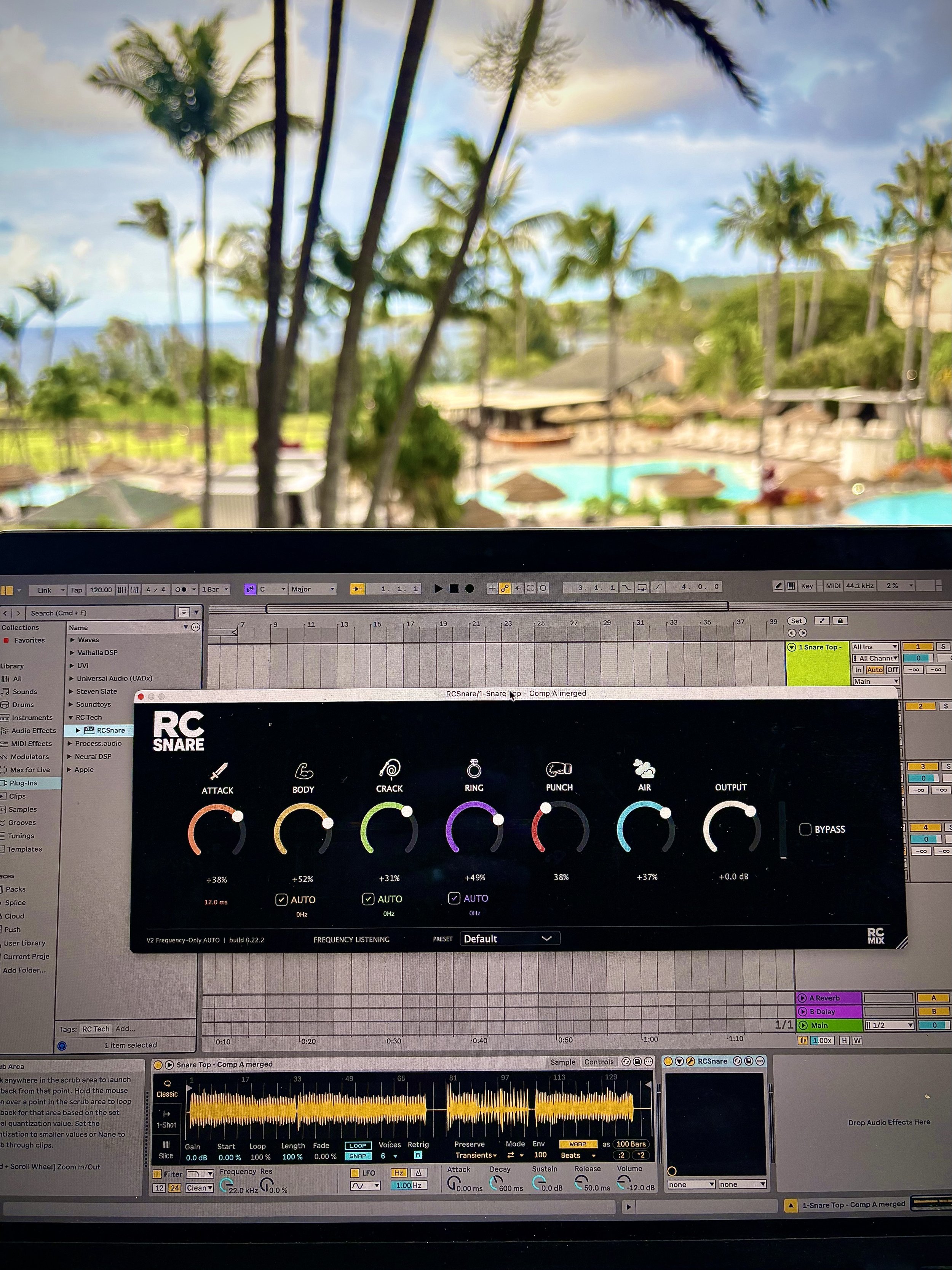 A computer screen showing music production software with various panels, including a drum machine interface labeled 'RC SNARE', waveforms, and track controls. The background view through a window depicts a tropical scene with palm trees and a swimming pool.
