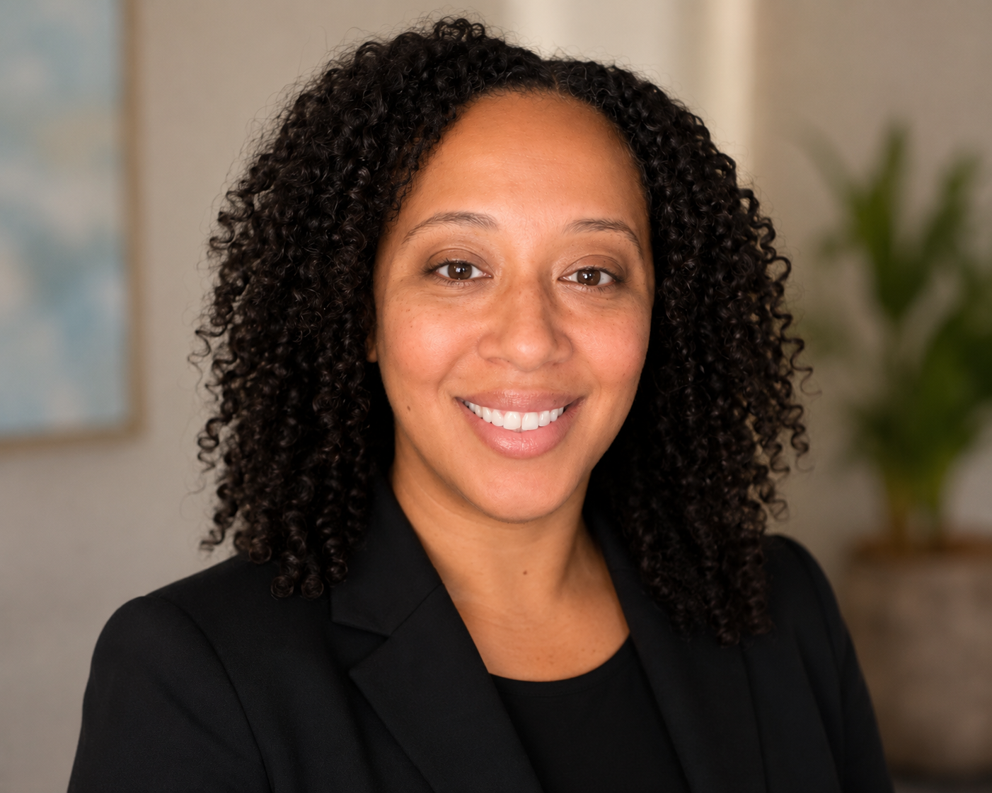 A professional woman with curly hair, wearing a black blazer, smiling in an indoor setting with a plant in the background.