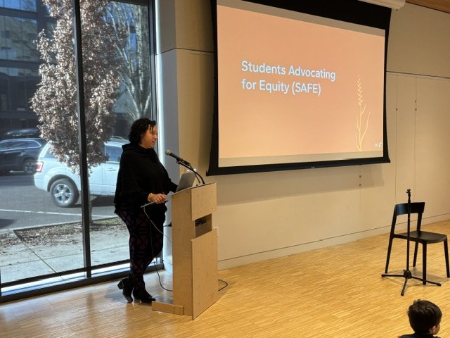 A woman standing at a podium speaking into a microphone during a presentation on 'Students Advocating for Equity (SAFE)' in a room with large windows and a projector screen.