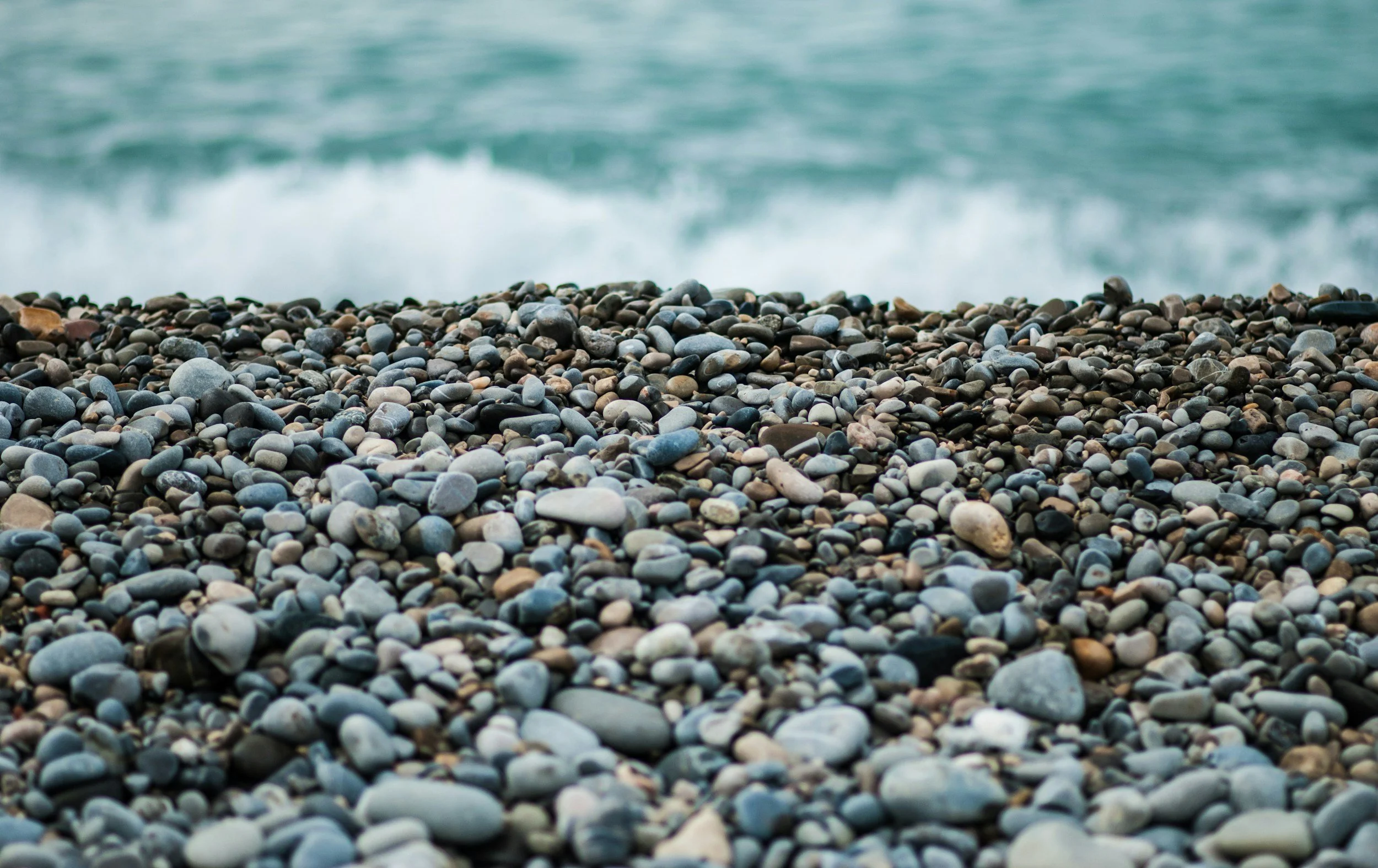 Pebble beach with small, multicolored round stones in the foreground and the ocean with waves in the background.
