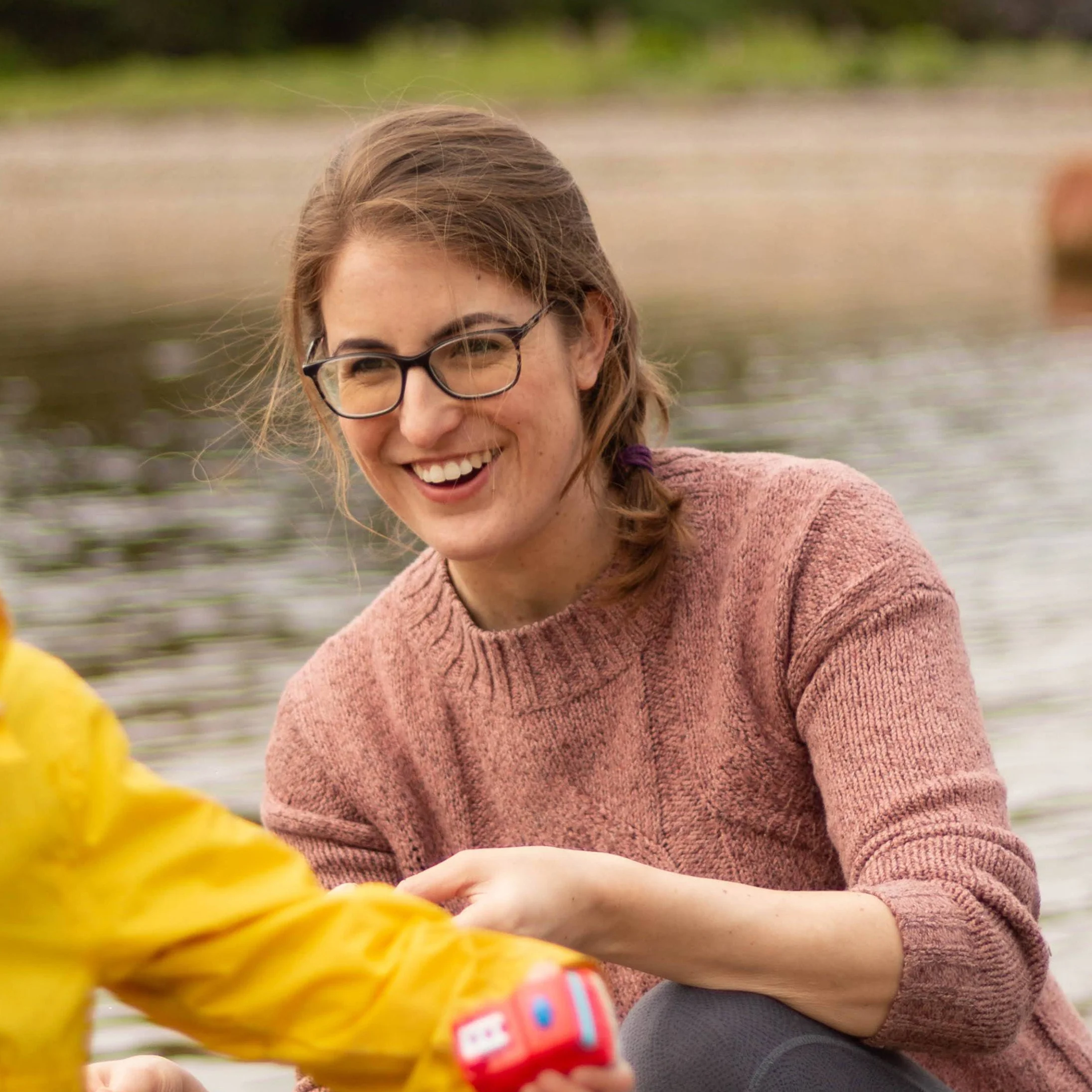 A smiling woman with glasses and braided hair wearing a pink sweater sitting outside near a body of water.