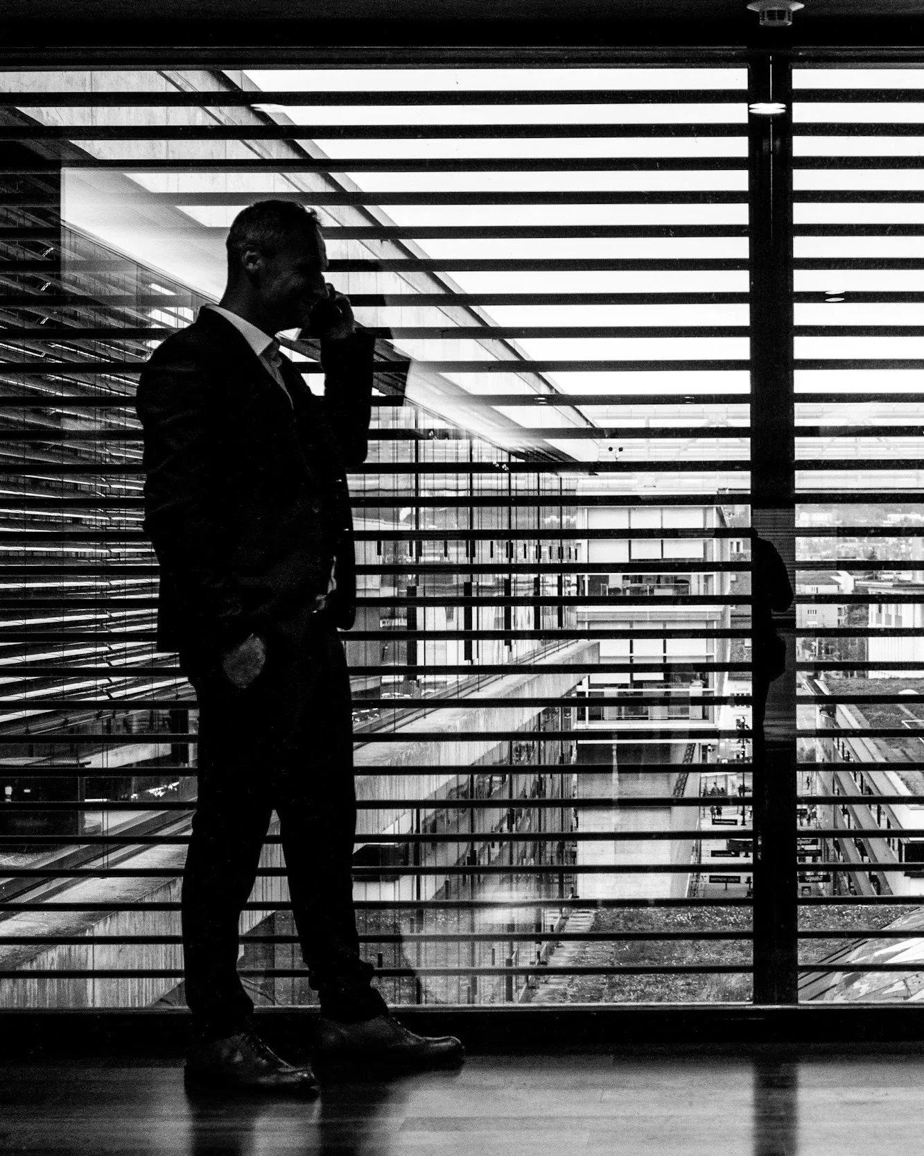 A silhouette of a man in a suit with a mobile phone to his ear, standing in front of a large window with horizontal blinds, overlooking a cityscape.