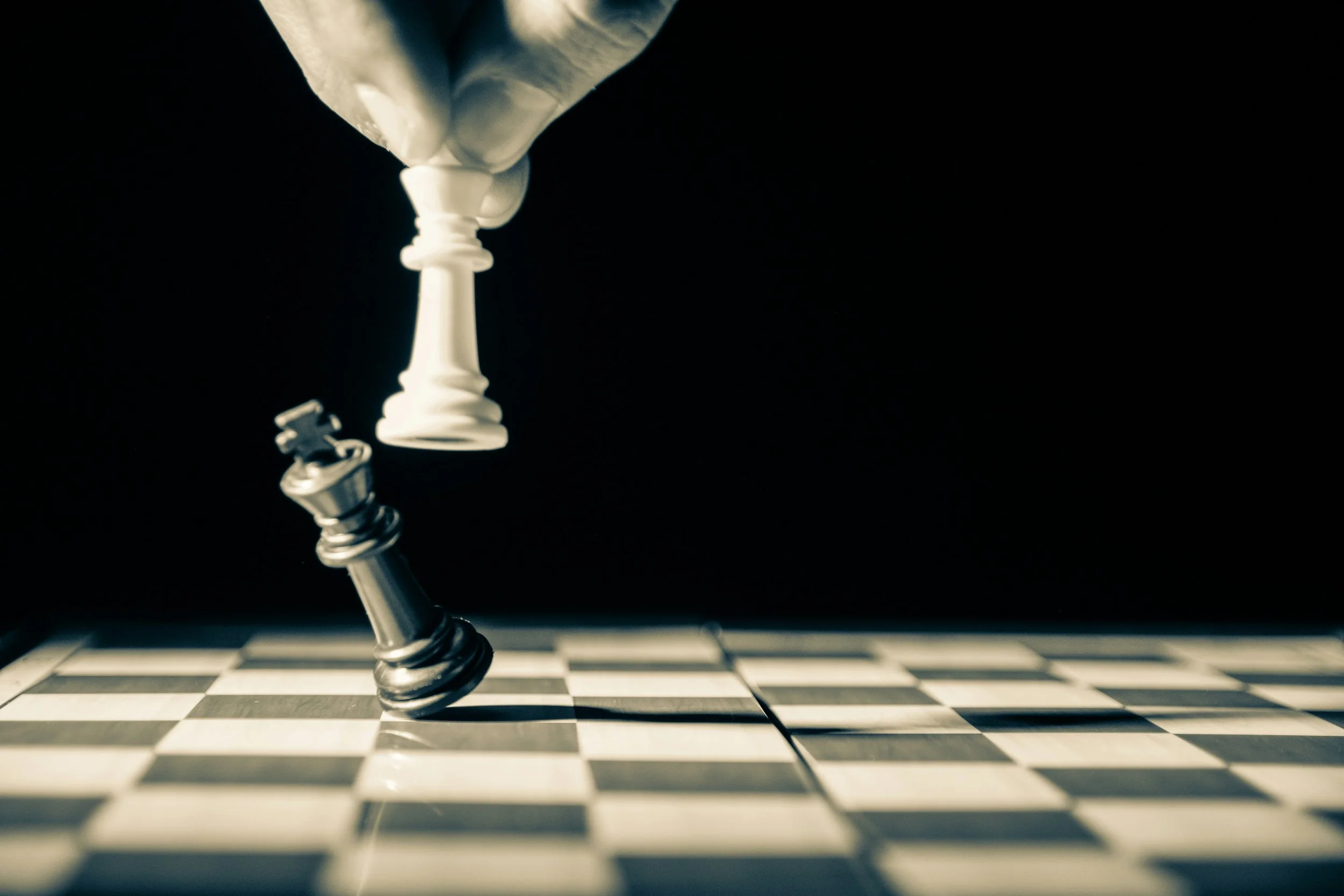 A close-up of a white king chess piece knocked over on a black and white checkered chessboard, with a black king chess piece lying on the side, against a dark background.