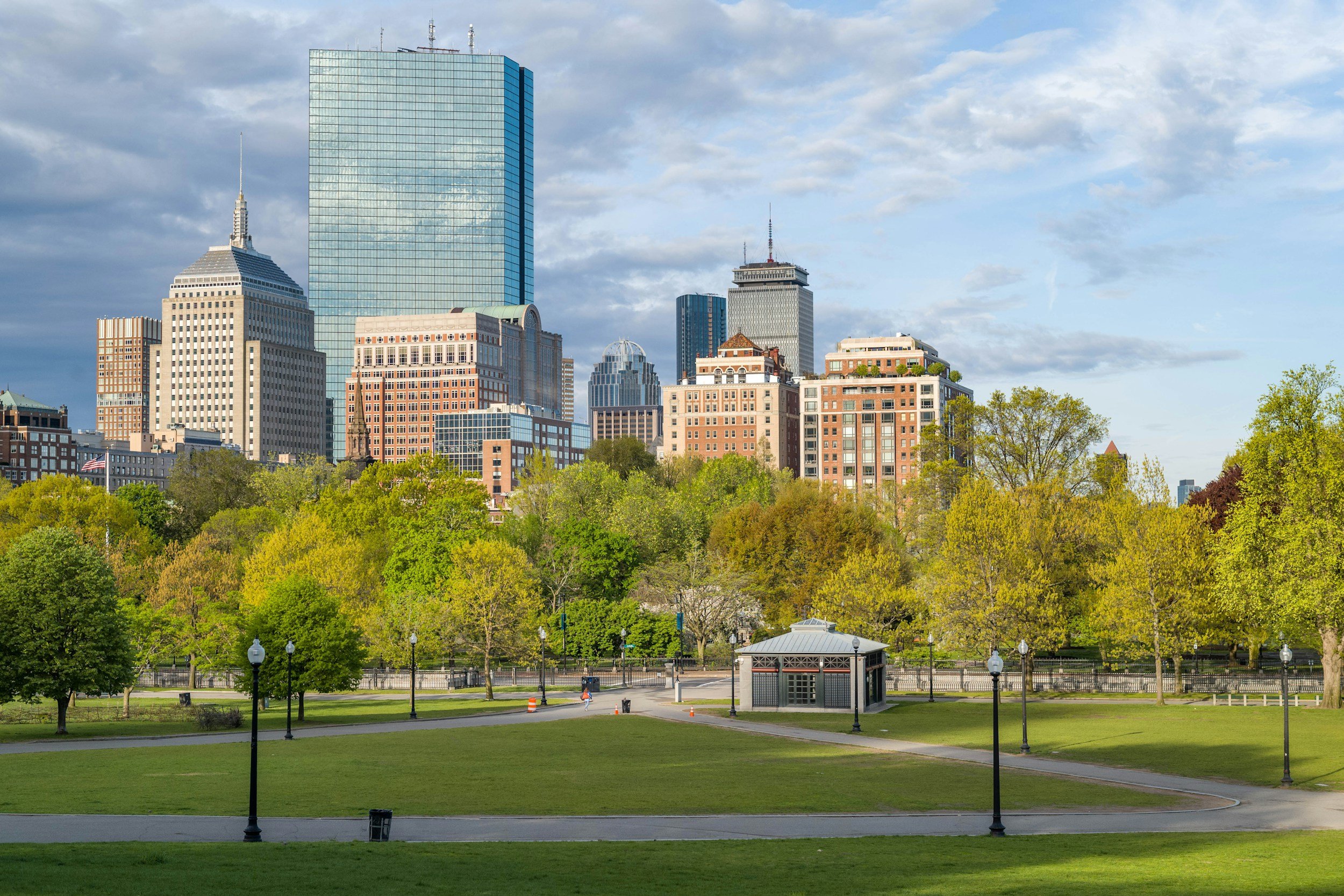 A park with green grass and trees, a small pavilion, street lamps, and a few people walking. In the background, a city skyline with high-rise buildings under a partly cloudy sky.