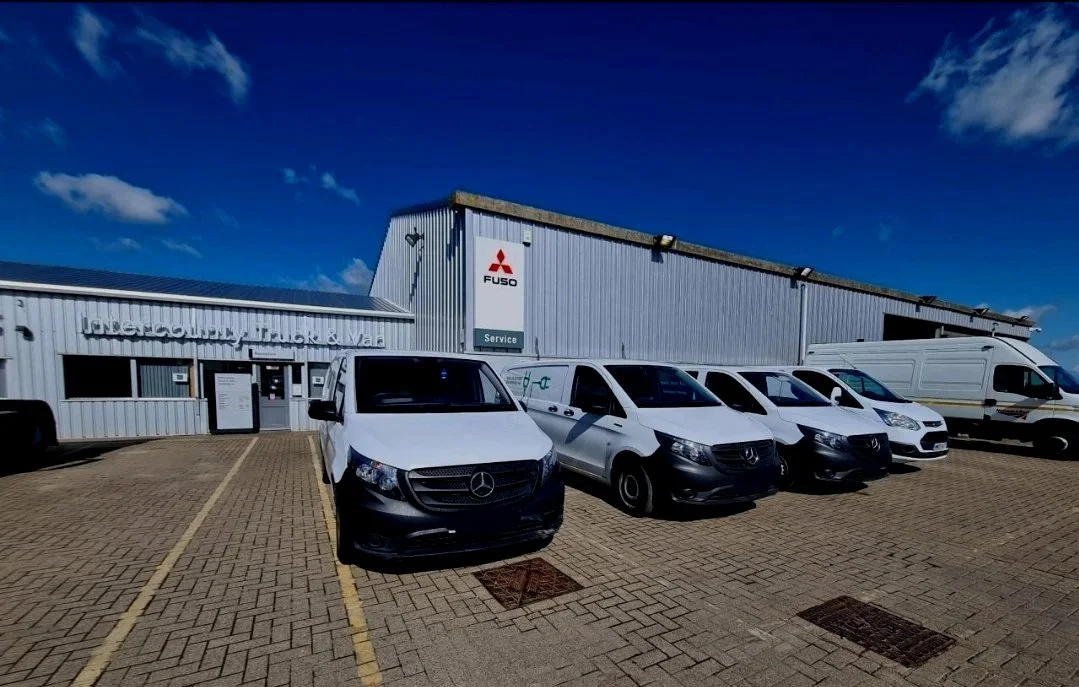 Parking lot with five white service vehicles in front of a building, labeled 'Intercounty Truck & Van' and 'Fuso'.