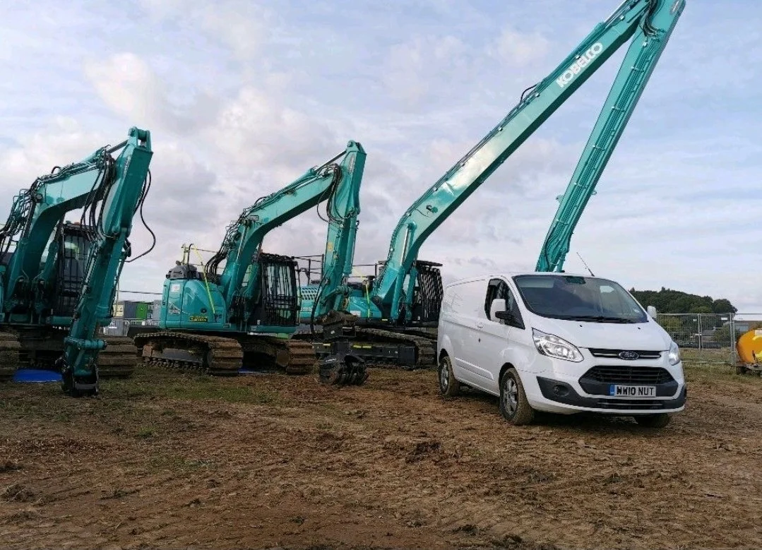 A white Ford Transit van parked next to three green excavators with extended arms on a dirt lot.