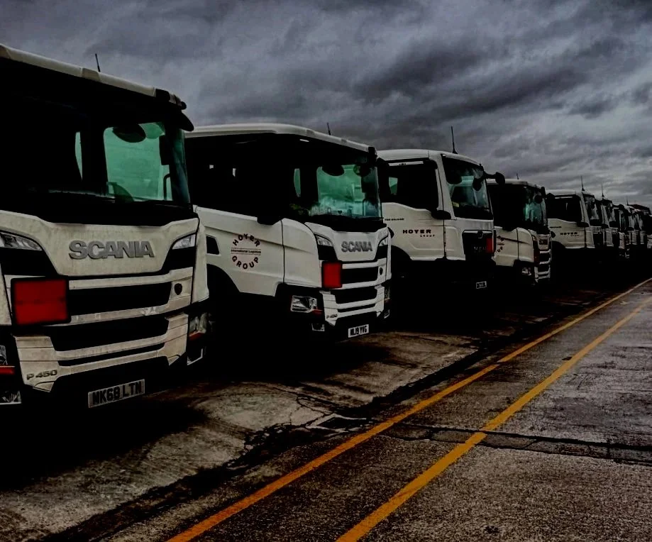 Lineup of large white trucks parked on a wet asphalt lot under dark, cloudy sky.