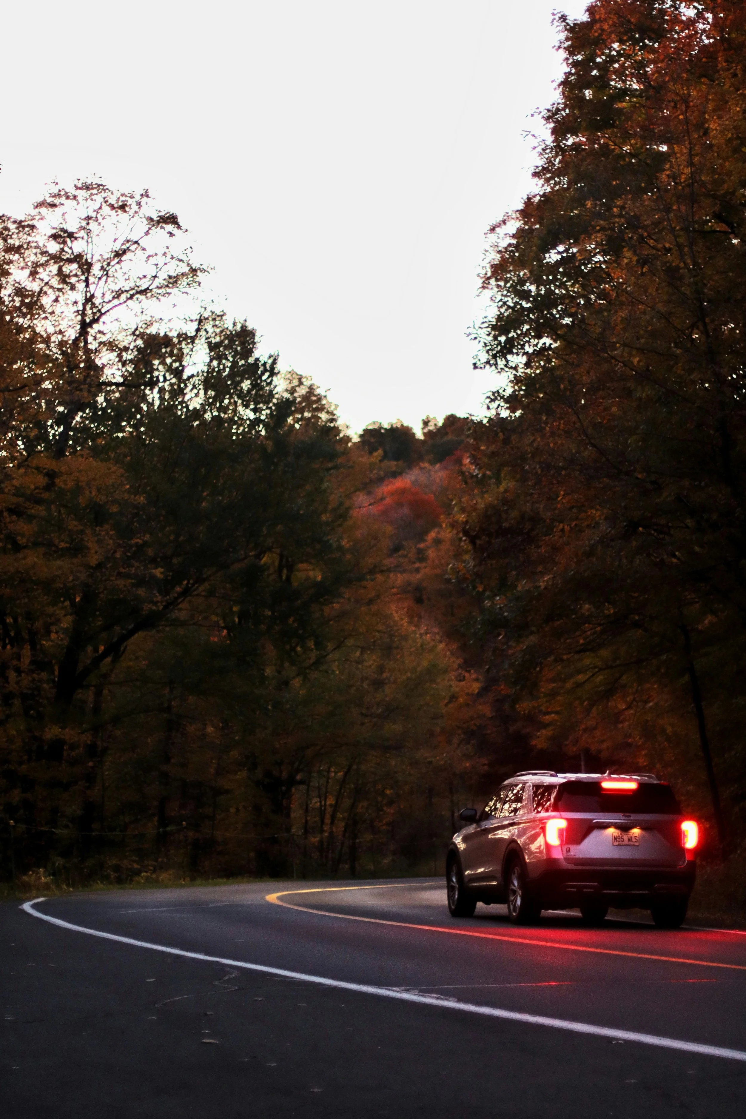 A black SUV driving on a winding road at dusk, surrounded by trees with fall foliage, with the sky mostly clear and the vehicle's brake lights illuminated.