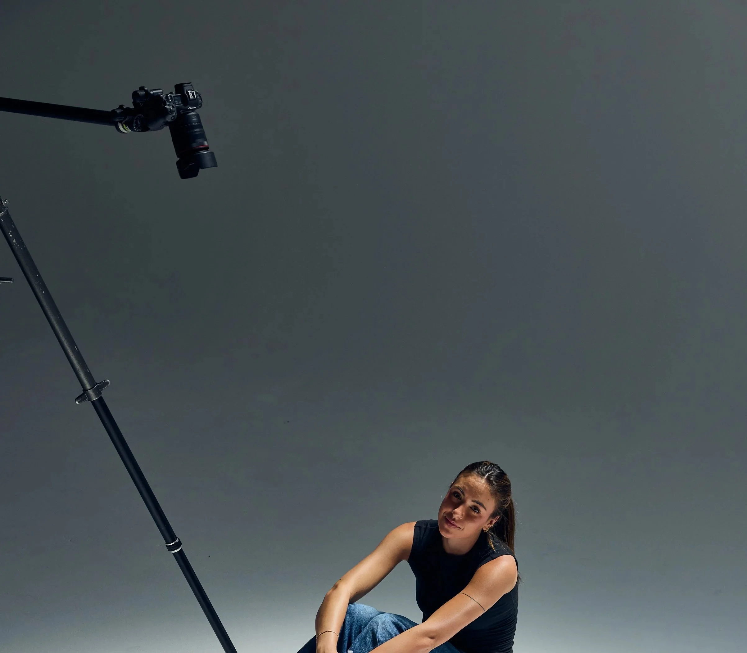 A woman with dark hair tied back, wearing a black shirt and blue jeans, is crouching down and smiling at the camera against a plain gray background. A camera on a tripod is positioned to the left of her, pointing towards her.