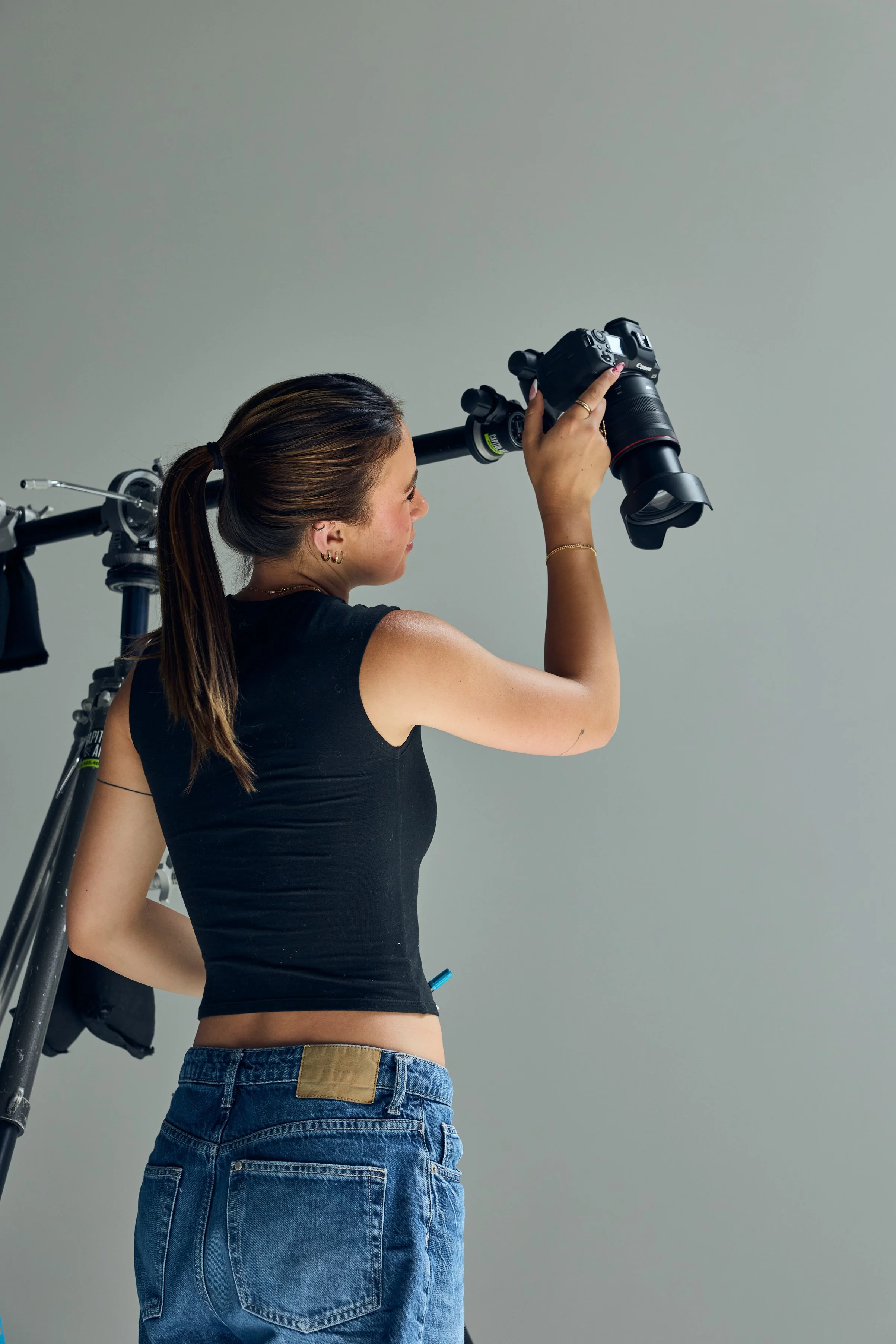 A woman holding a professional camera with a telephoto lens, standing next to a camera stabilizer against a plain, neutral background.