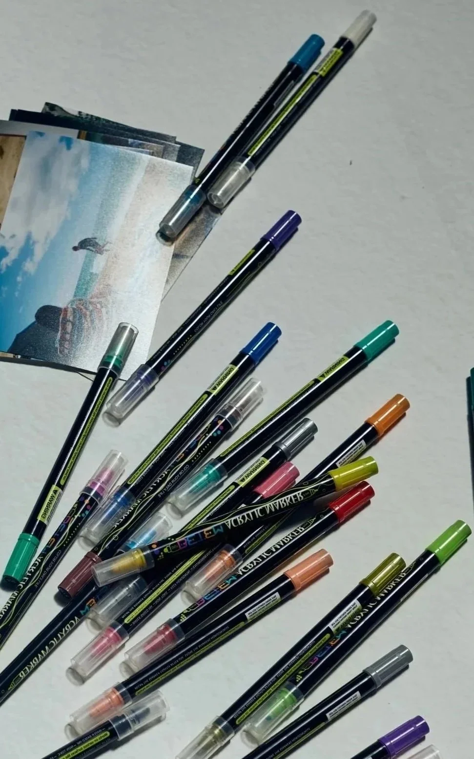 A collection of colorful brush tip markers scattered on a white surface, with some markers uncapped and a few small printed photos nearby.
