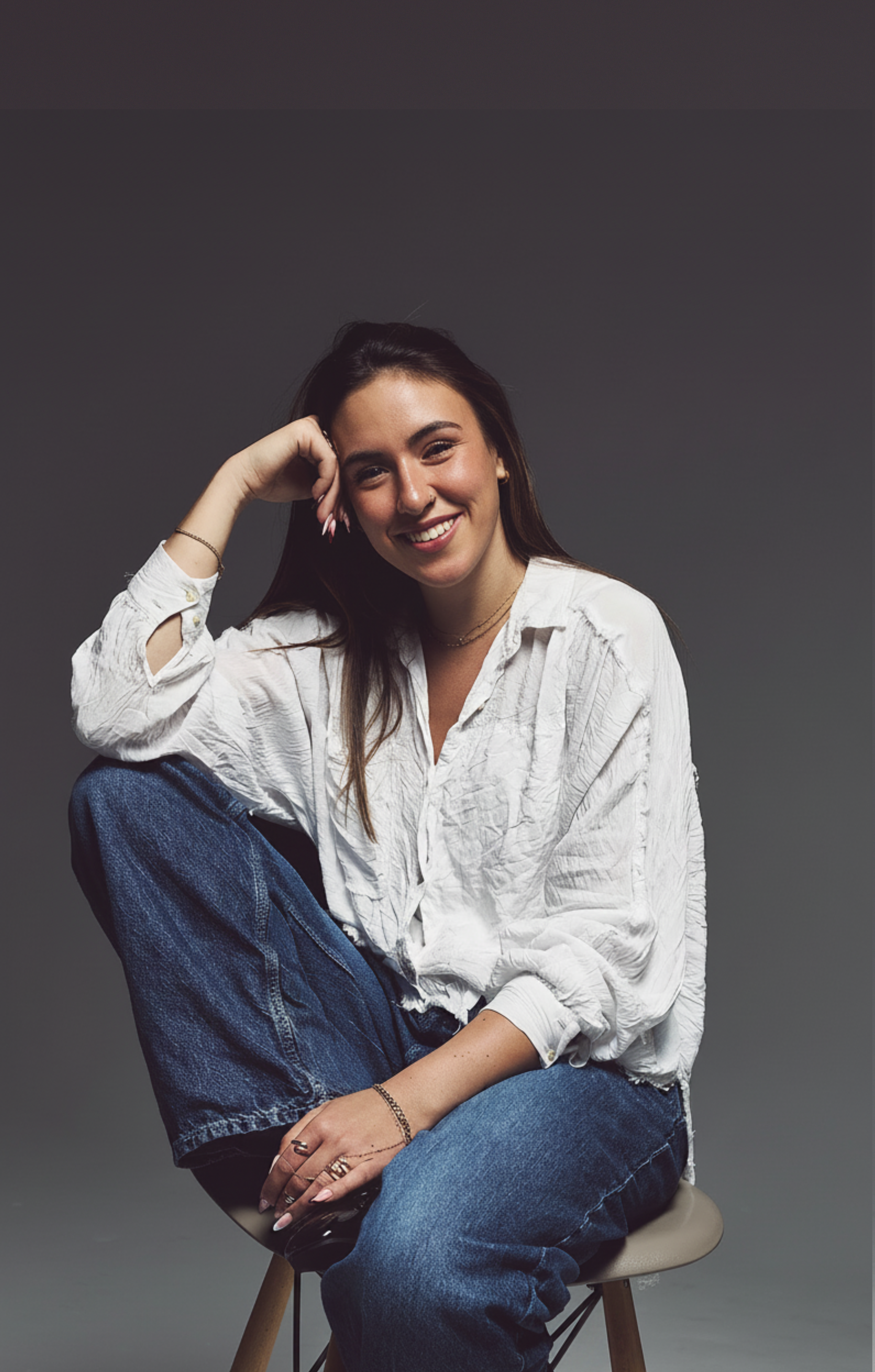 A woman sitting on a stool, smiling at the camera, with her left hand on her head and dressed in a white, crinkled shirt and blue jeans.