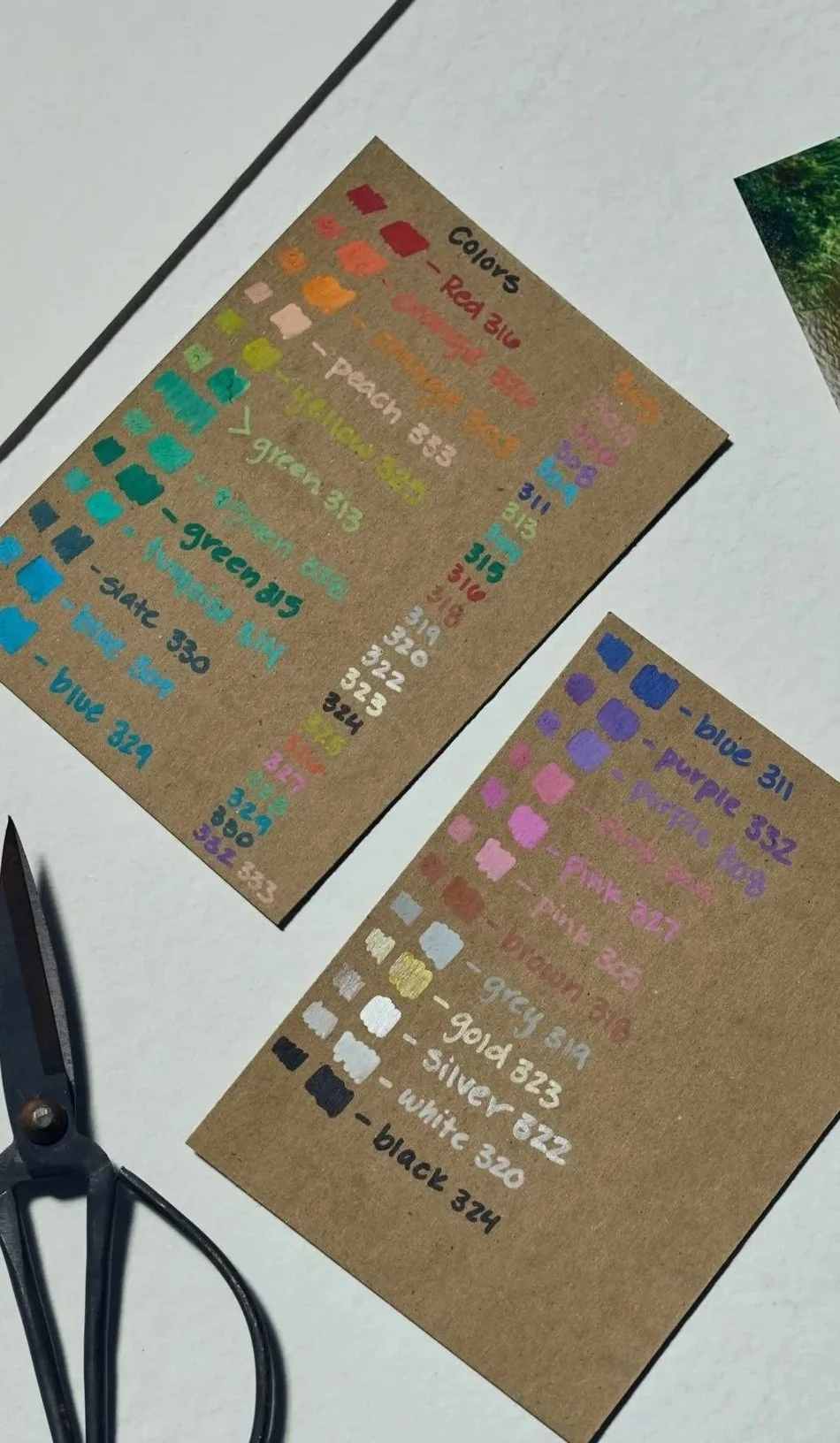 Color chart on brown paper with various shades of paint, each labeled with color name and code, including greens, pinks, blues, purples, and neutrals.