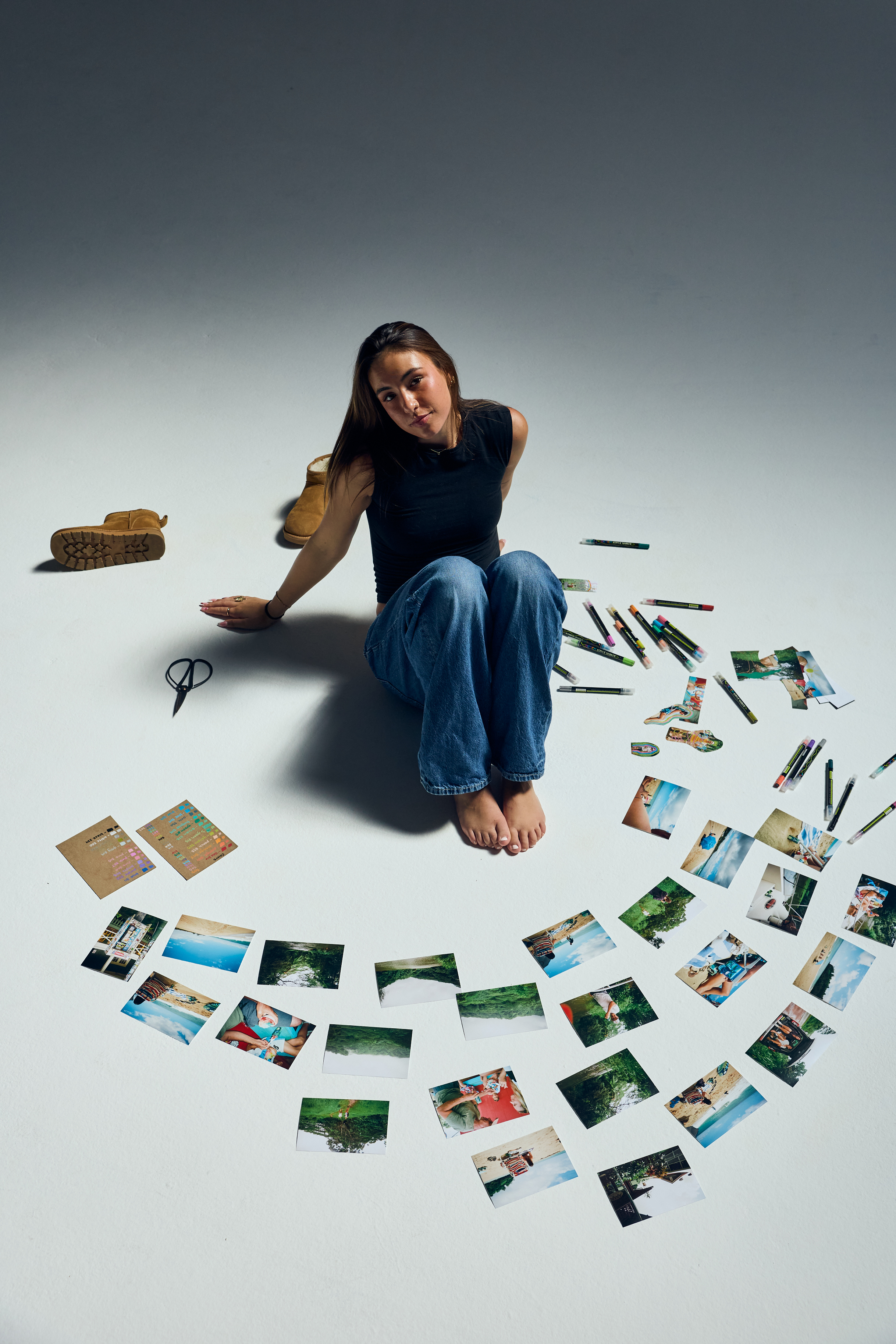 A young woman with long brown hair, wearing a black sleeveless top and blue jeans, sitting on the floor in a photo studio. She is surrounded by scattered printed photographs and art supplies, including markers, scissors, and photographs. She is looking directly at the camera with a slight smile.