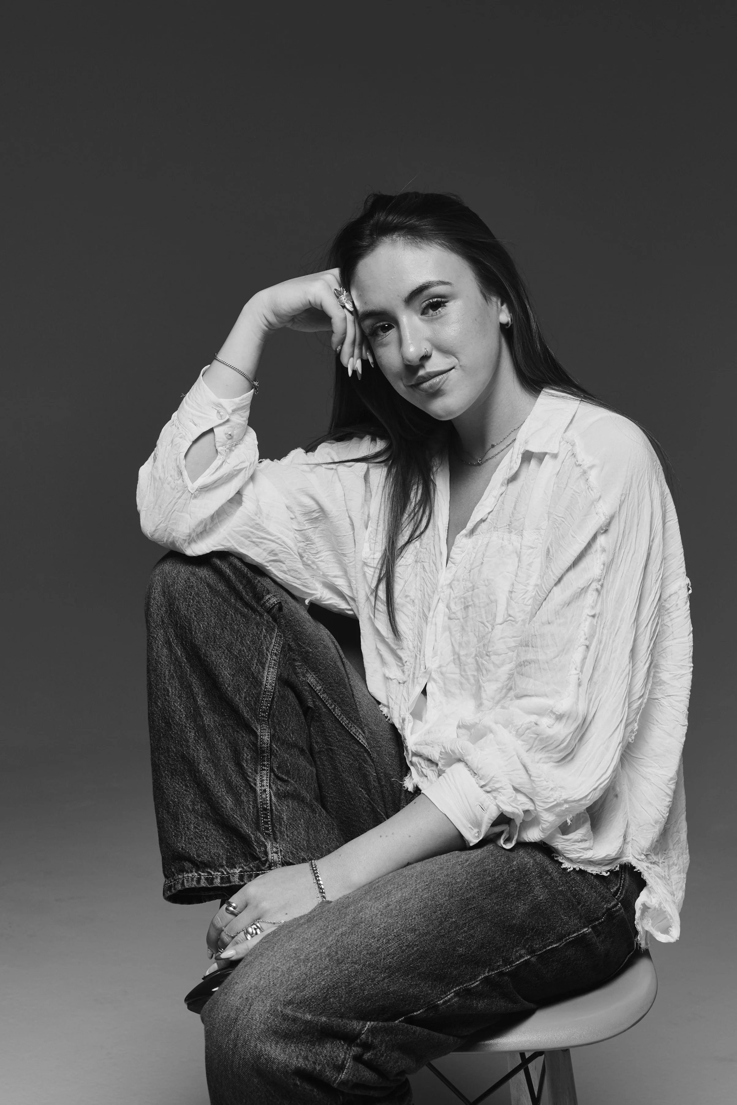 A black and white photo of a young woman sitting on a stool, looking at the camera with a slight smile. She has long dark hair, a nose piercing, and is wearing a light-colored, crinkled shirt with rolled up sleeves, dark jeans, and various jewelry including rings, bracelets, and necklaces. She is resting her head on her left hand with her elbow on her knee, and her right hand rests on her thigh.