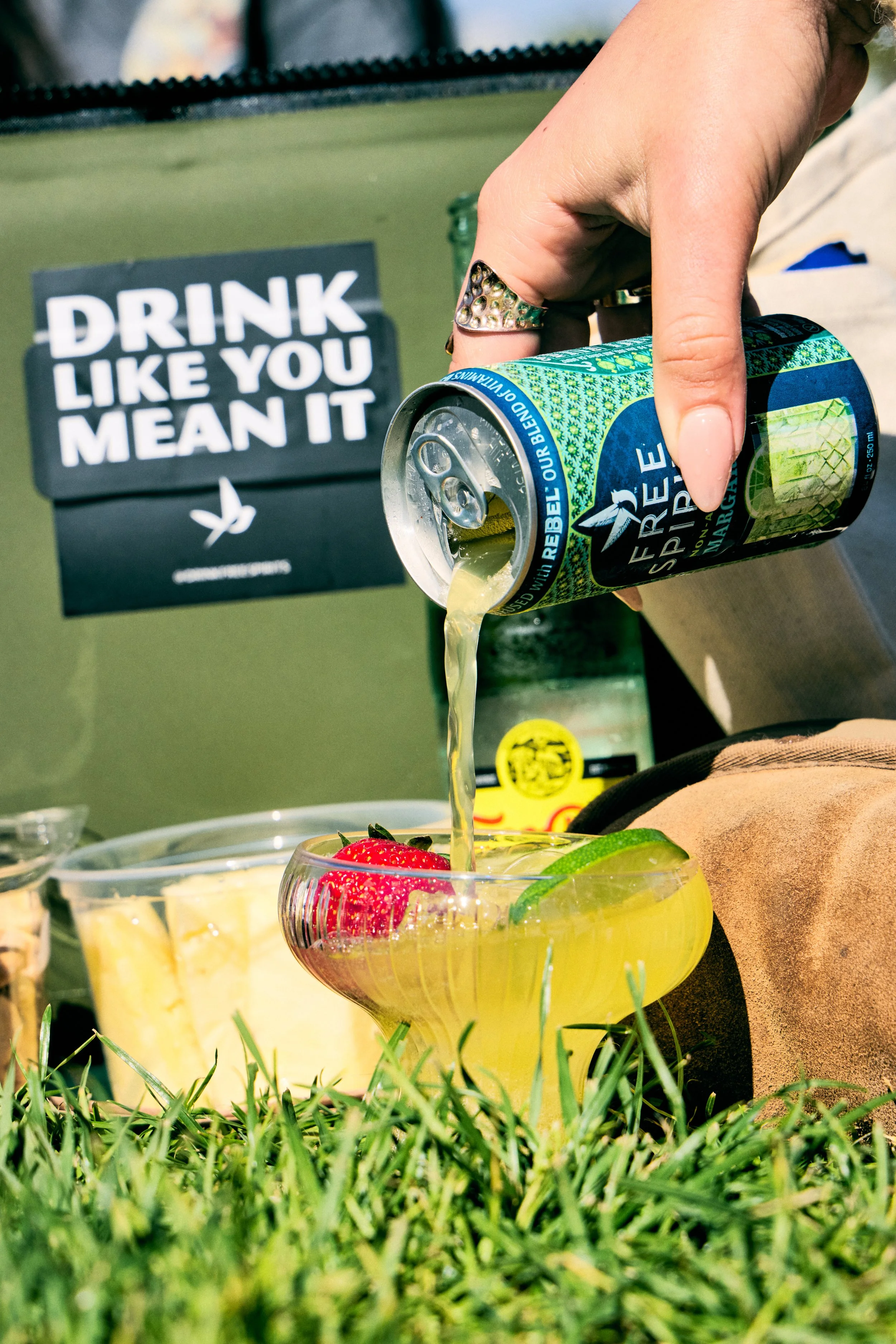 A person pouring a sparkling water drink into a cocktail glass with strawberries and lime slices on top, with a sign in the background that reads 'Drink Like You Mean It' and a display of fruit and drinks on a table, green grass at the foreground.