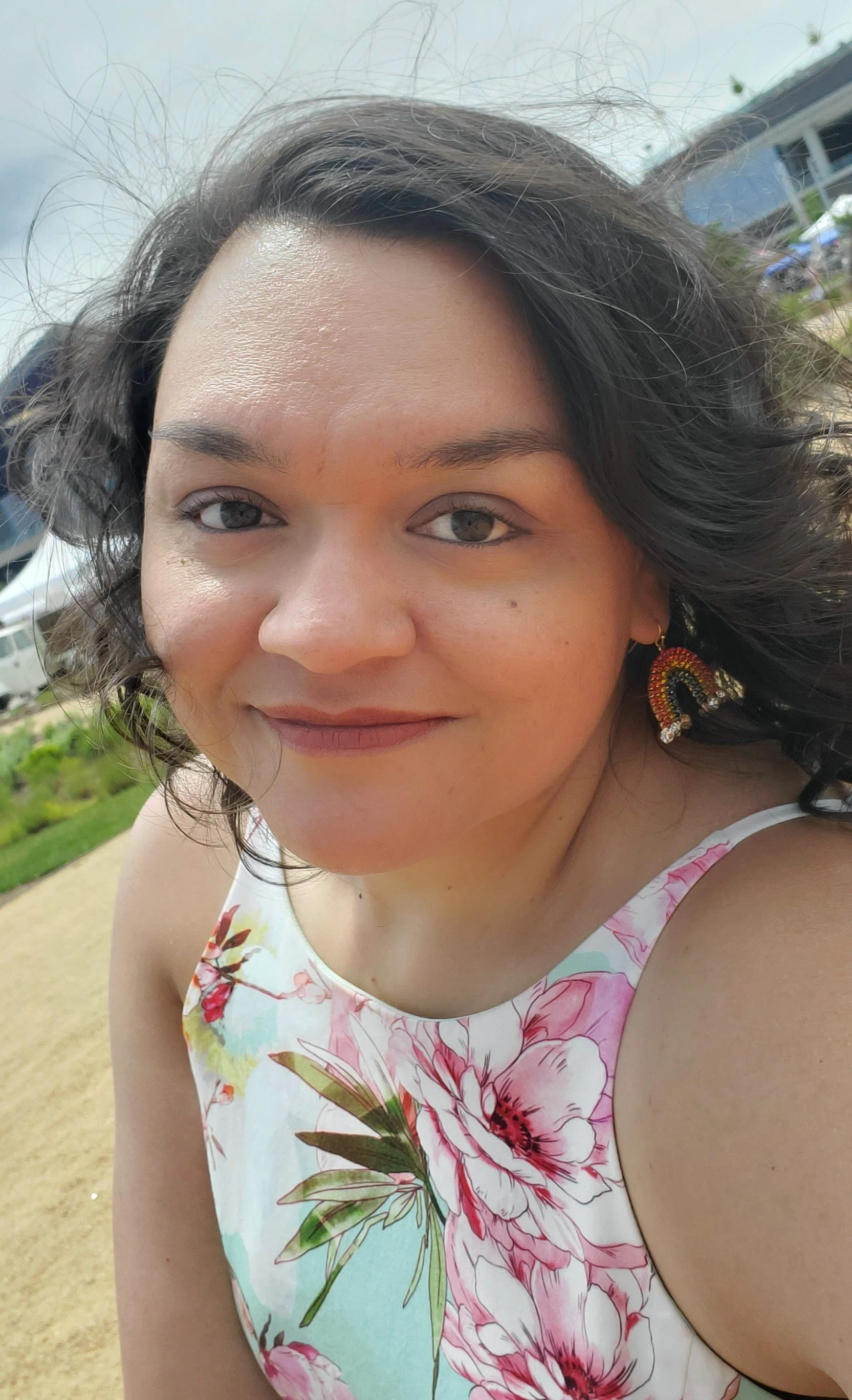 Smiling woman wearing rainbow earrings with windswept light curls in her hair on a partly cloudy day